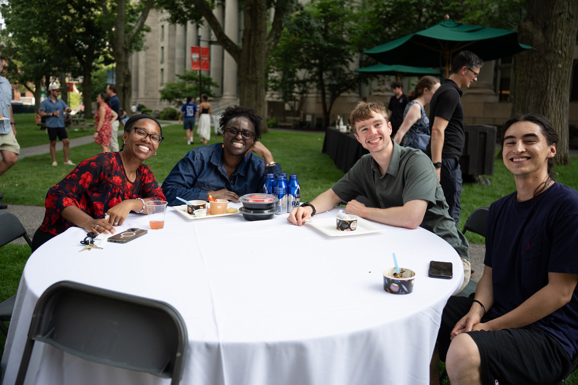 Four individuals smile while seated around a table outdoors, enjoying food and drinks with trees and other people in the background.
