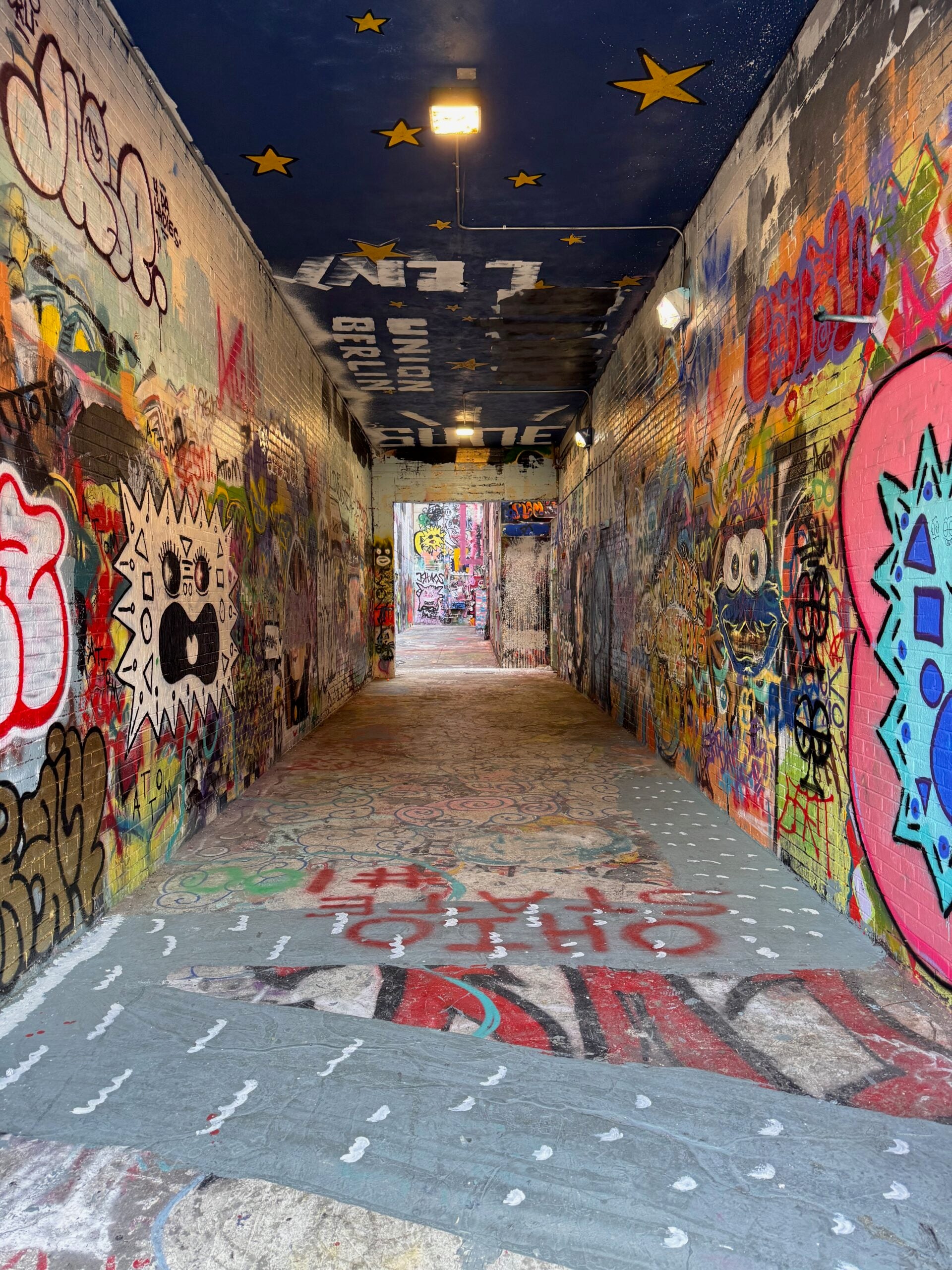 A graffiti-covered pedestrian tunnel in Ann Arbor, with colorful murals on the brick walls and yellow stars painted across the dark ceiling.