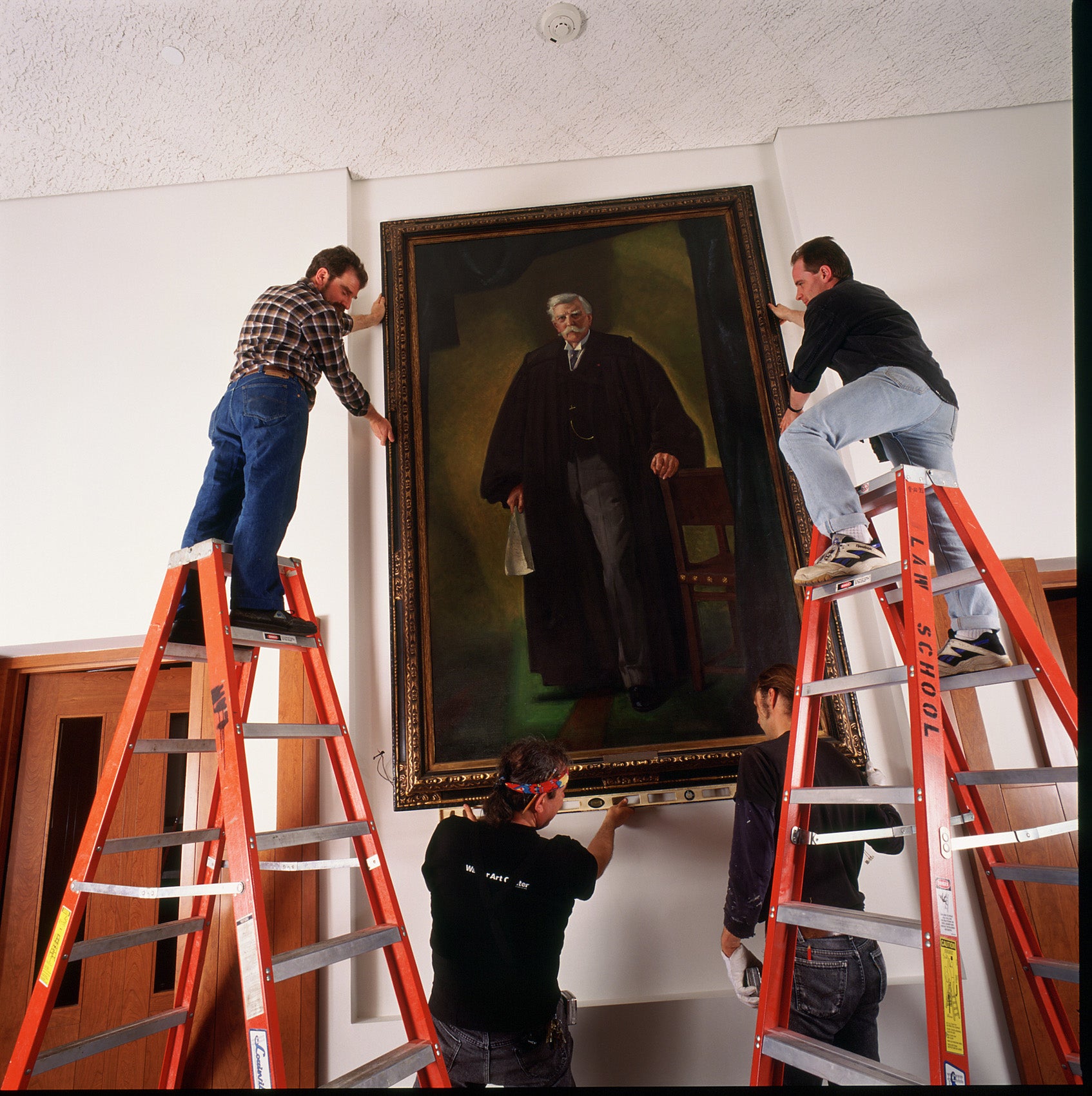 Two men on ladders install a large oil painting on a wall.