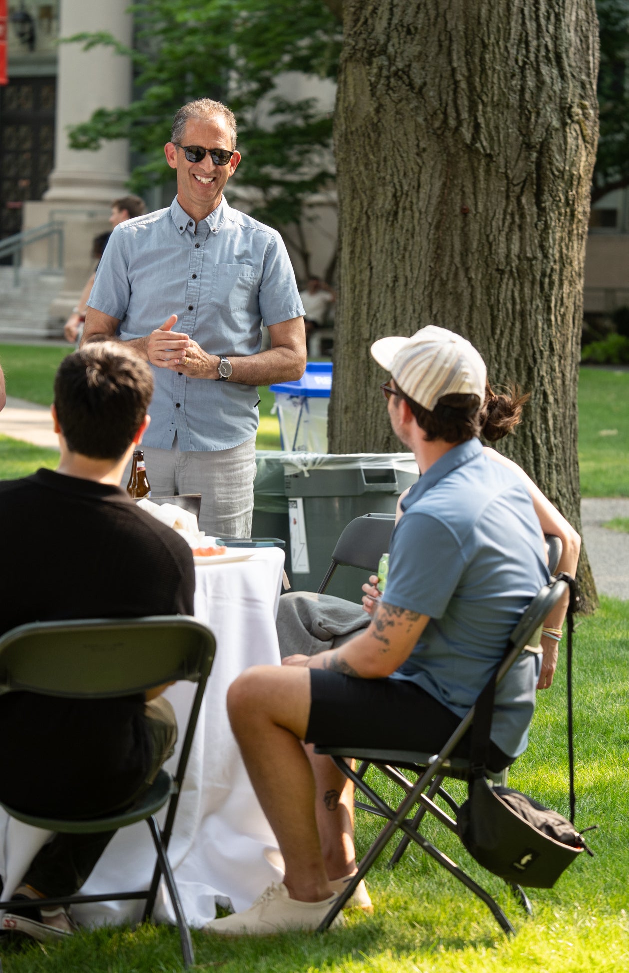 Several individuals are gathered outdoors around a table on a grassy lawn, engaged in conversation under a tree.