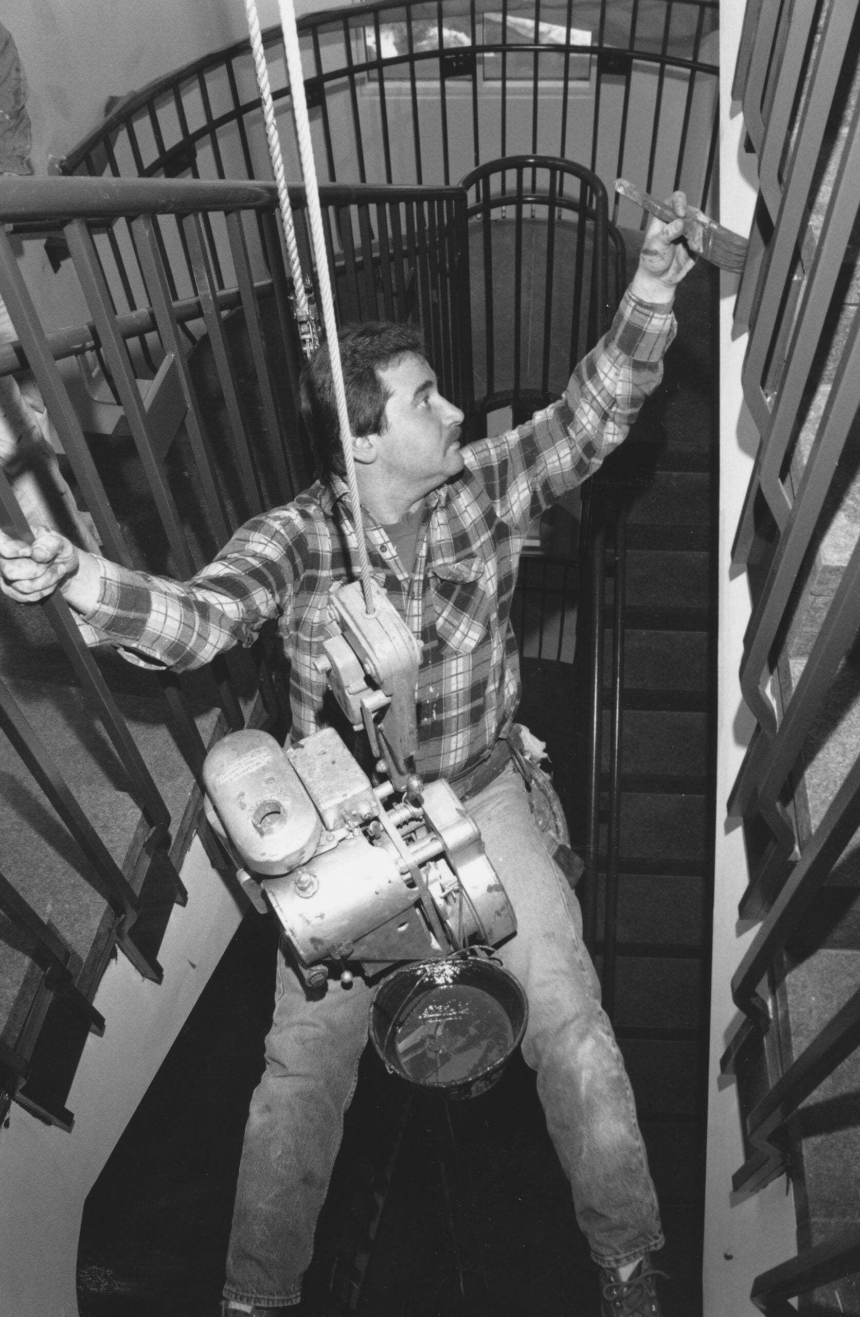 A painter, suspended from the ceiling, paints the railings on a staircase.
