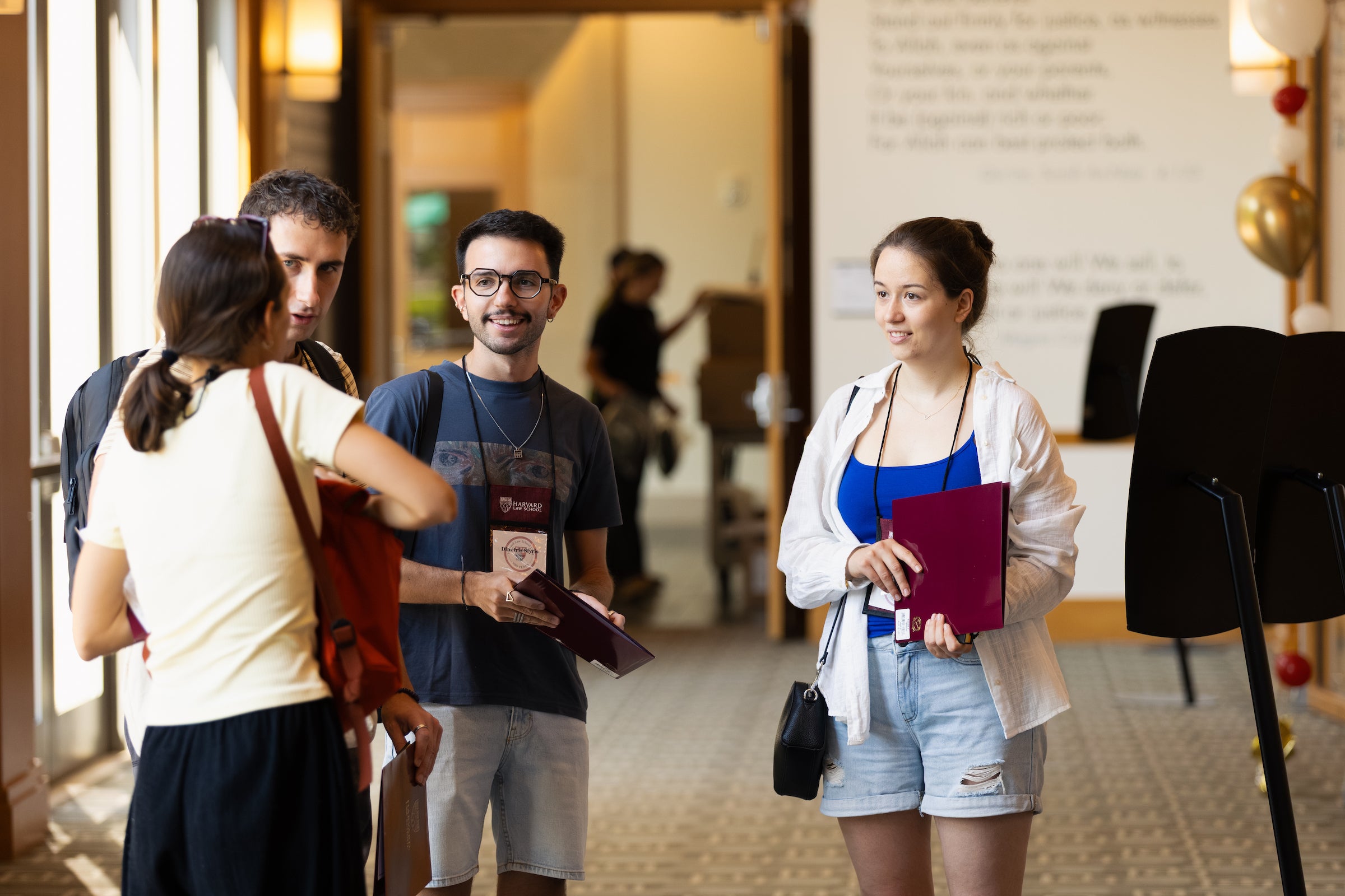 A group of students engage in conversation in a brightly lit indoor hallway, with two people holding folders and wearing lanyards.