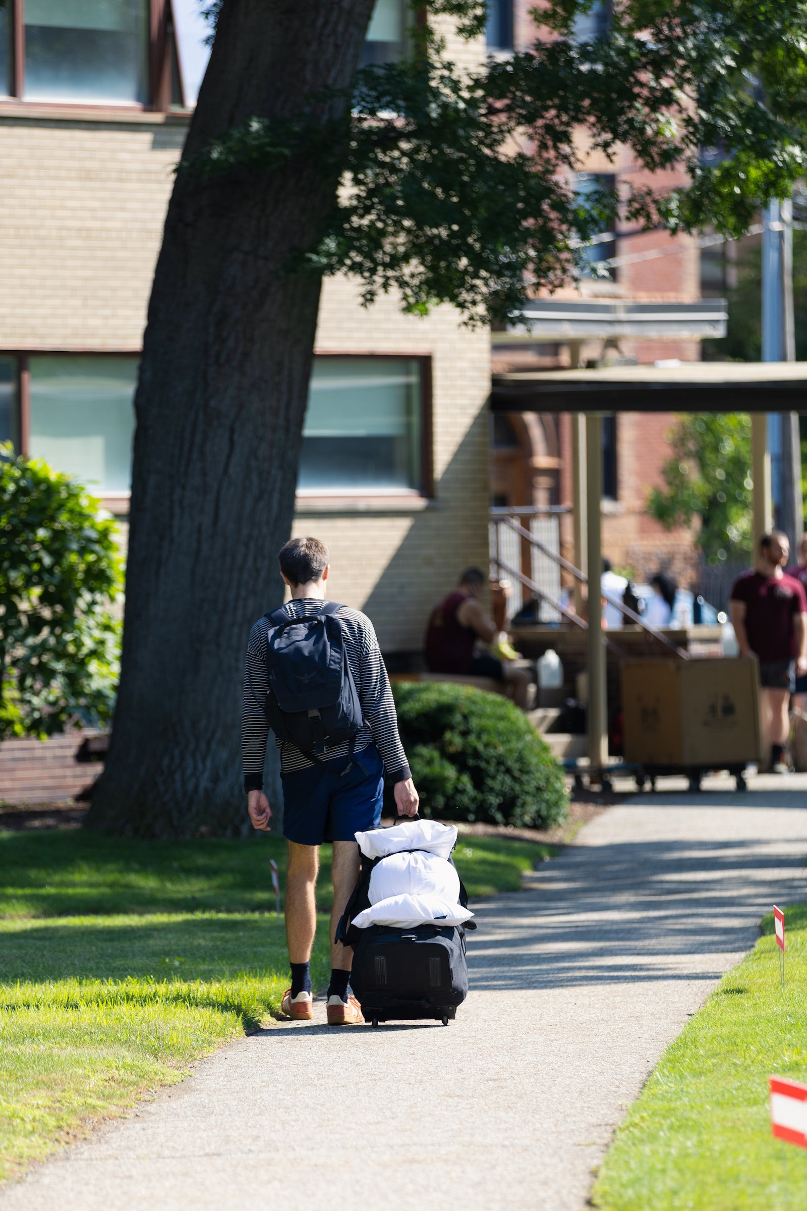 A person wearing a backpack walks along a paved path, pulling a suitcase loaded with a white pillow.