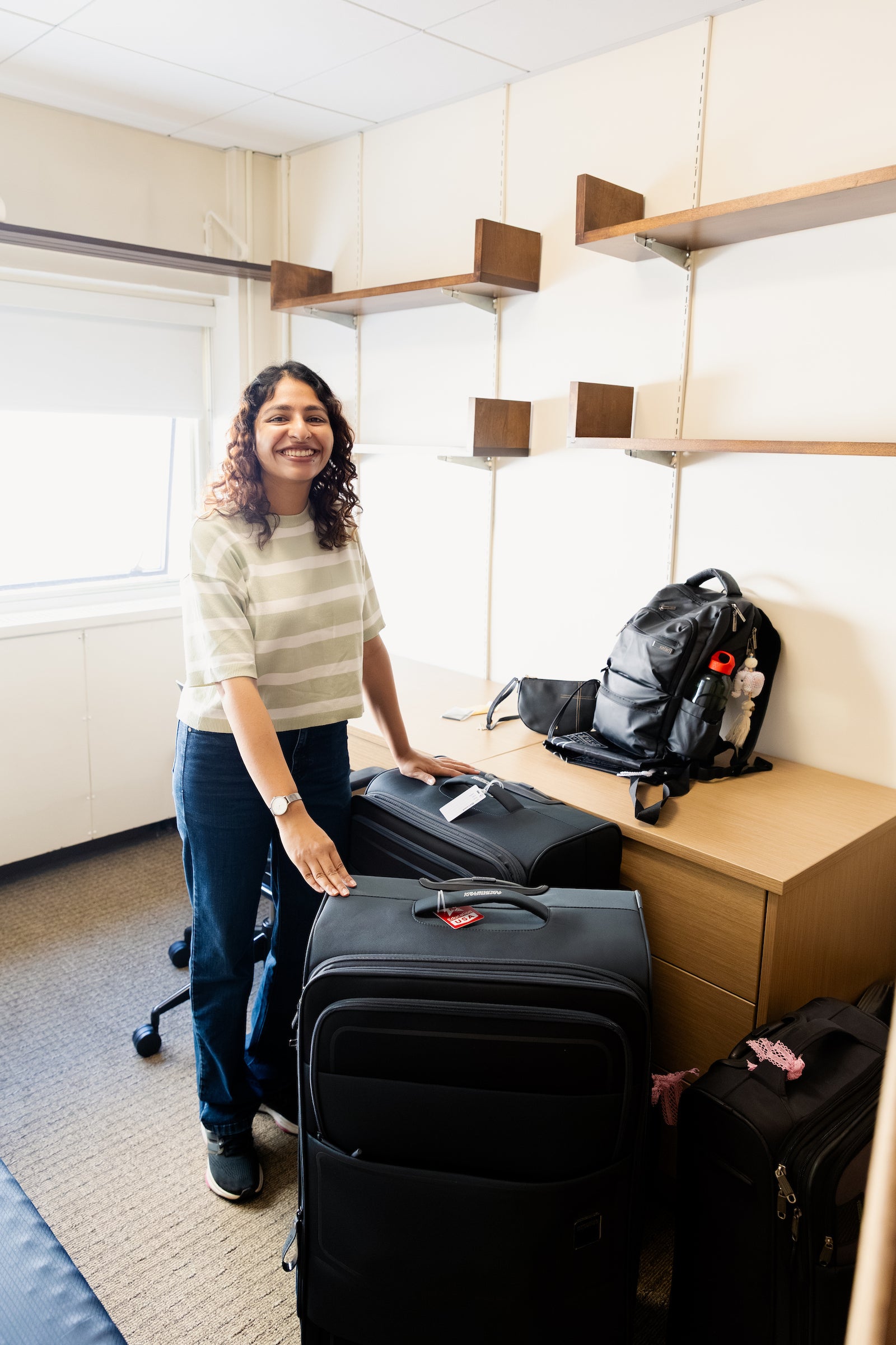 A student smiles while standing next to several suitcases in a room with wooden shelves and a desk.