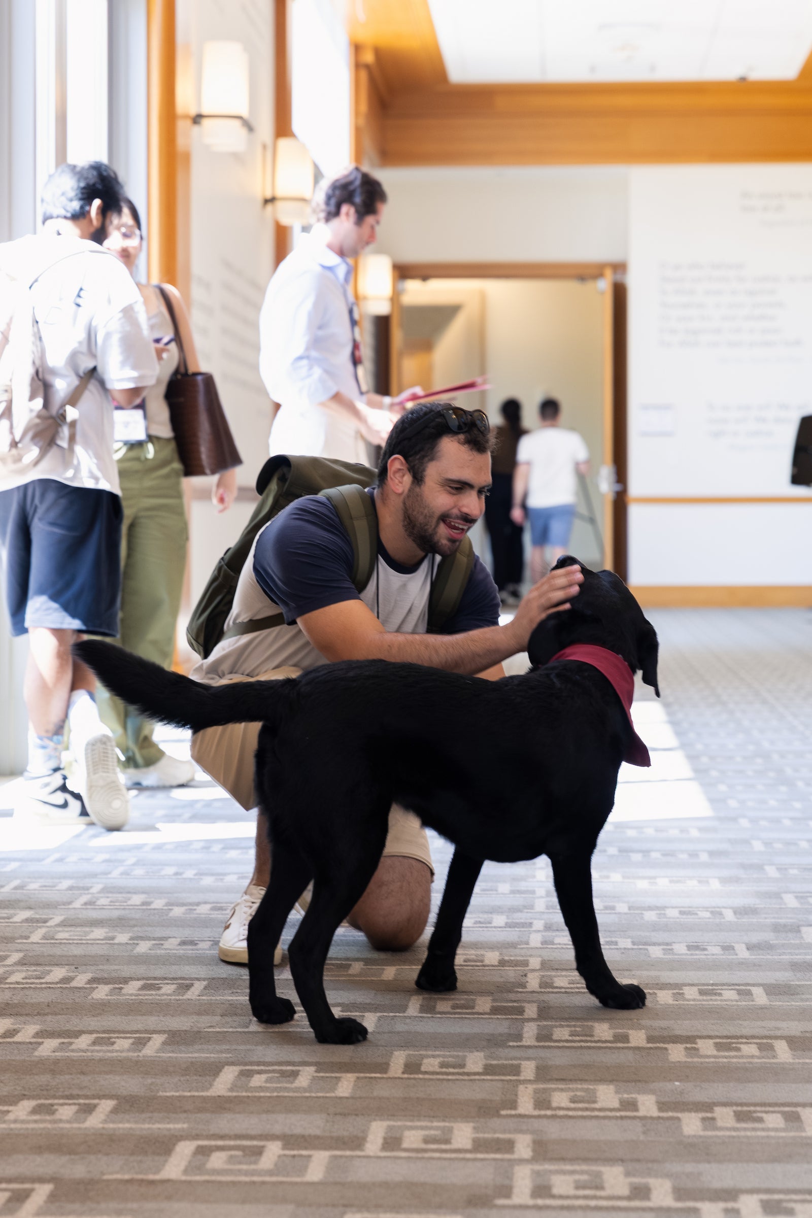 A student pets a black dog in a hallway with other students.