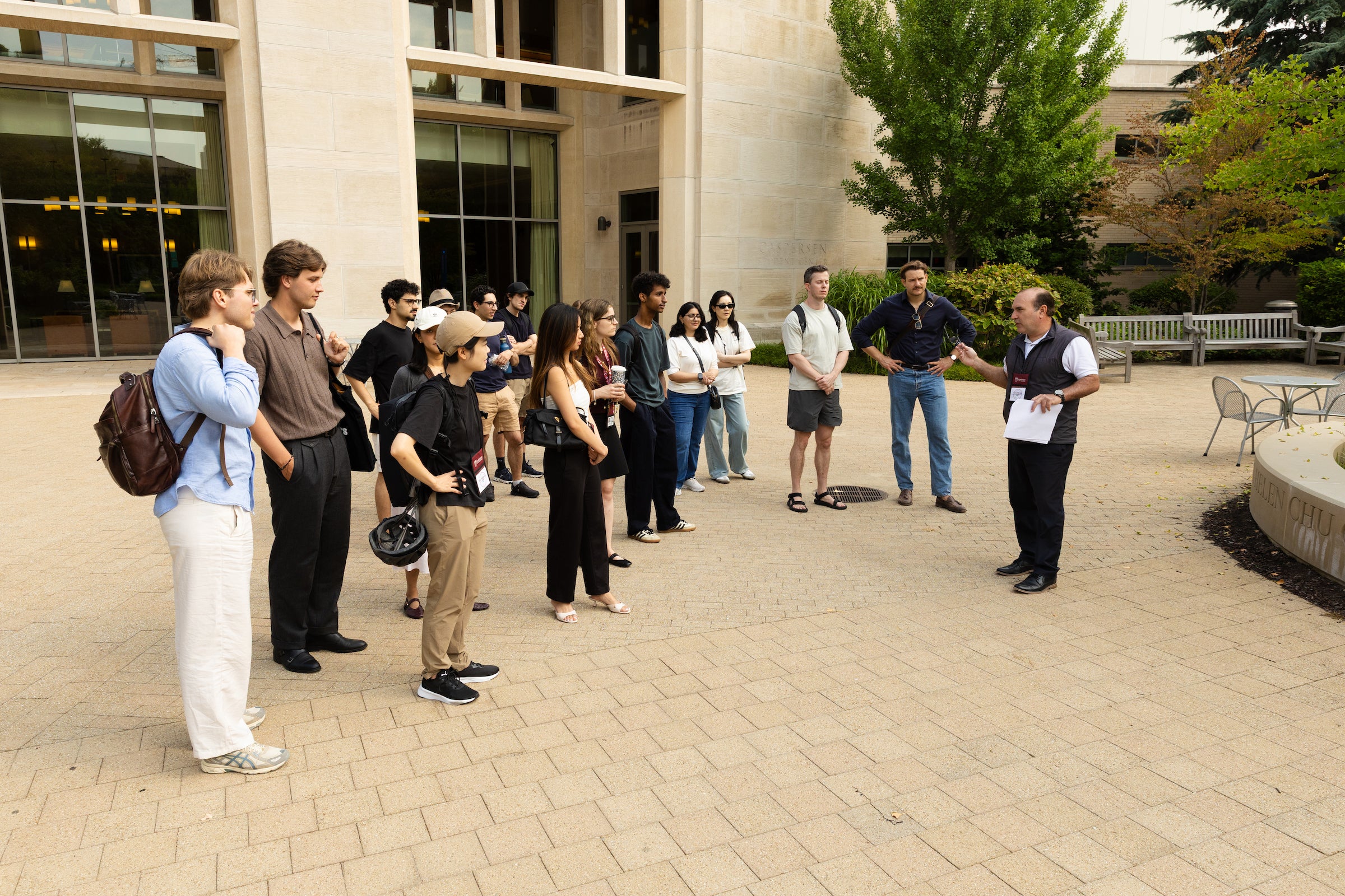 A group of students gather in a courtyard to listen to a guide who is speaking and gesturing while holding papers.