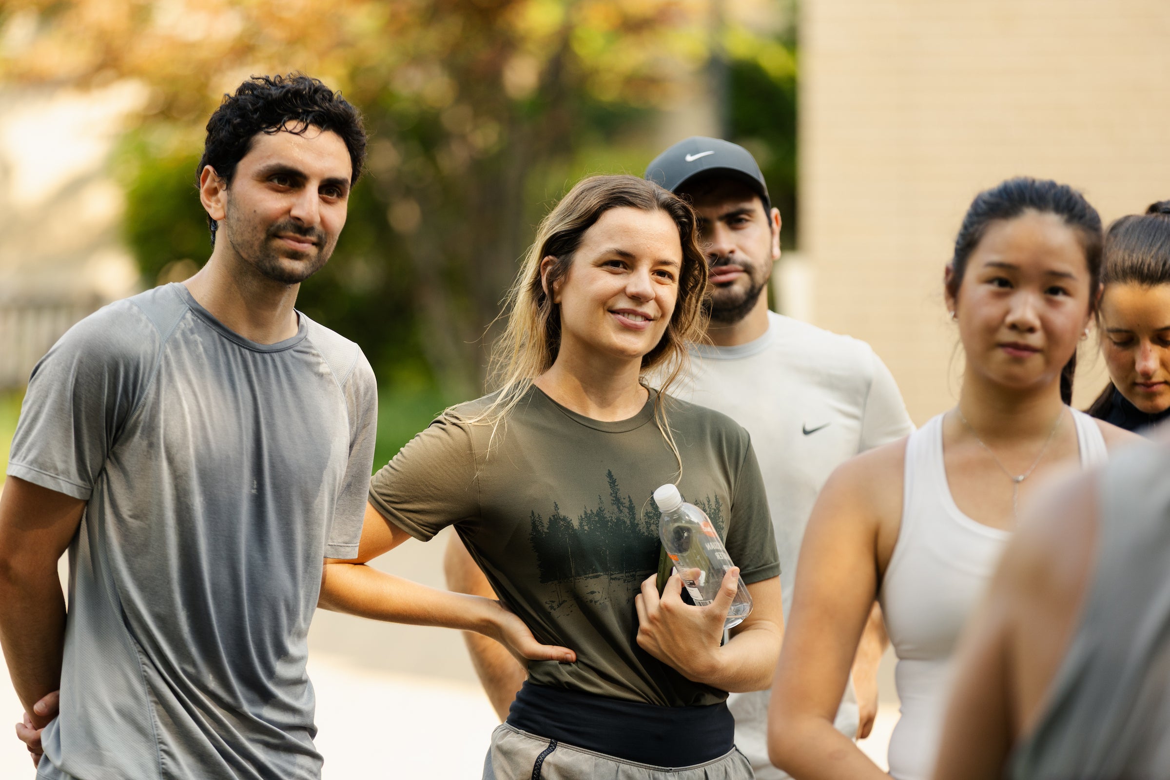 A group of people in athletic wear stand outdoors, with one person holding a water bottle and smiling at a speaker.
