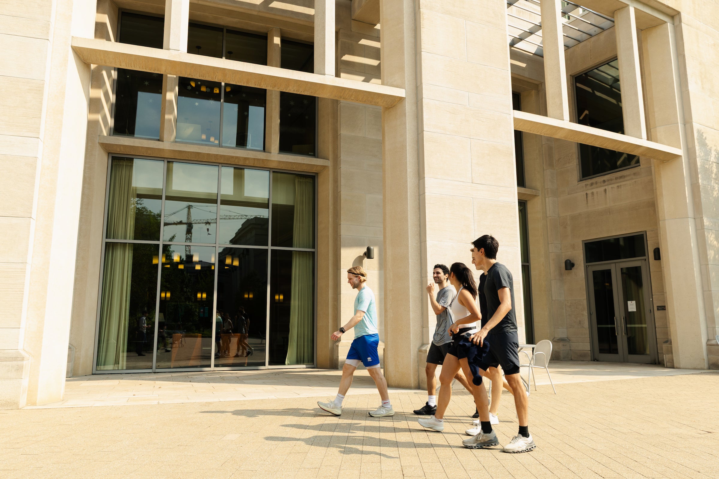 Four students walk across a sunlit plaza in front of a modern stone and glass building.