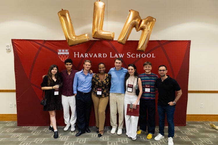 A group of eight students pose in front of a Harvard Law School banner with large gold “LLM” balloons above them.