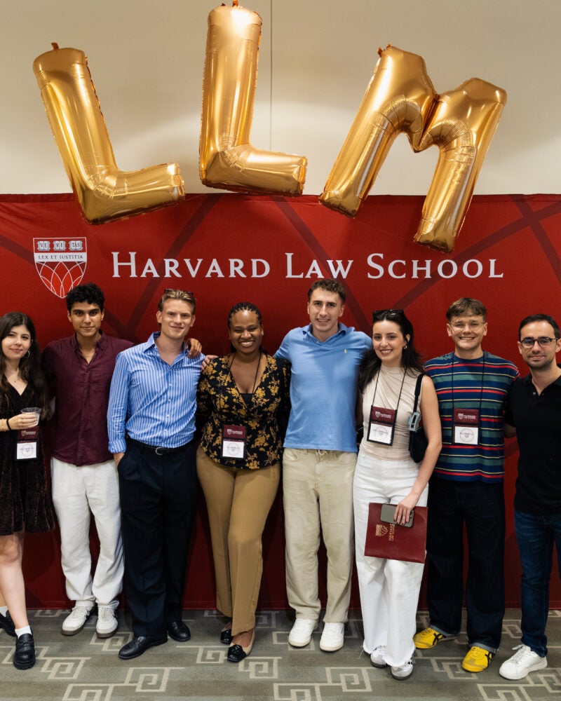 A group of eight students pose in front of a Harvard Law School banner with large gold “LLM” balloons above them.