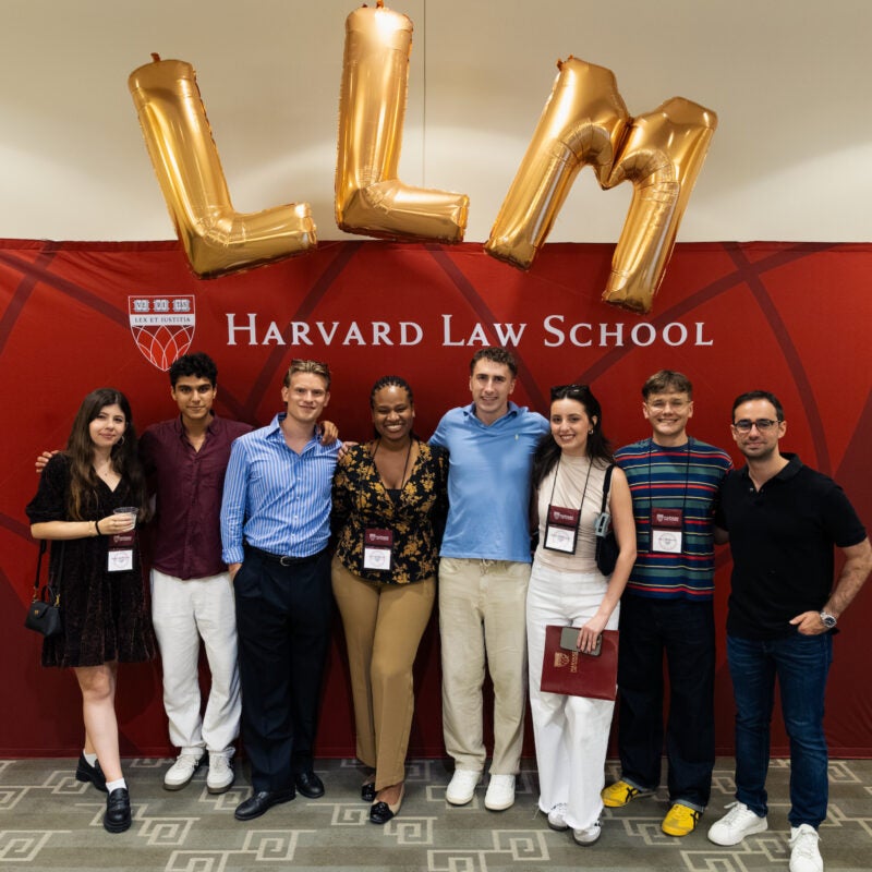 A group of eight students pose in front of a Harvard Law School banner with large gold “LLM” balloons above them.