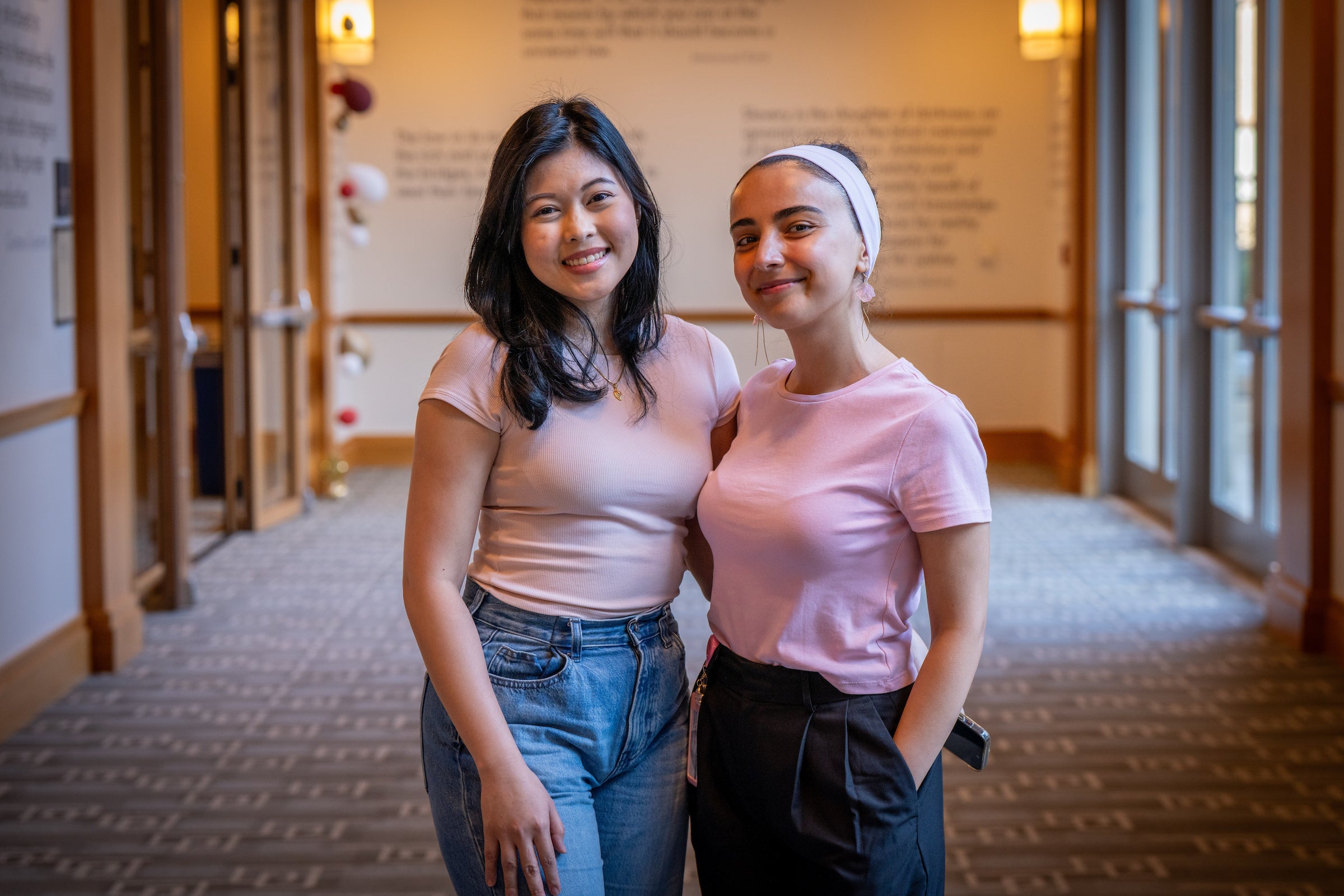 Two individuals in matching pink tops stand together in a warmly lit hallway, smiling at the camera.