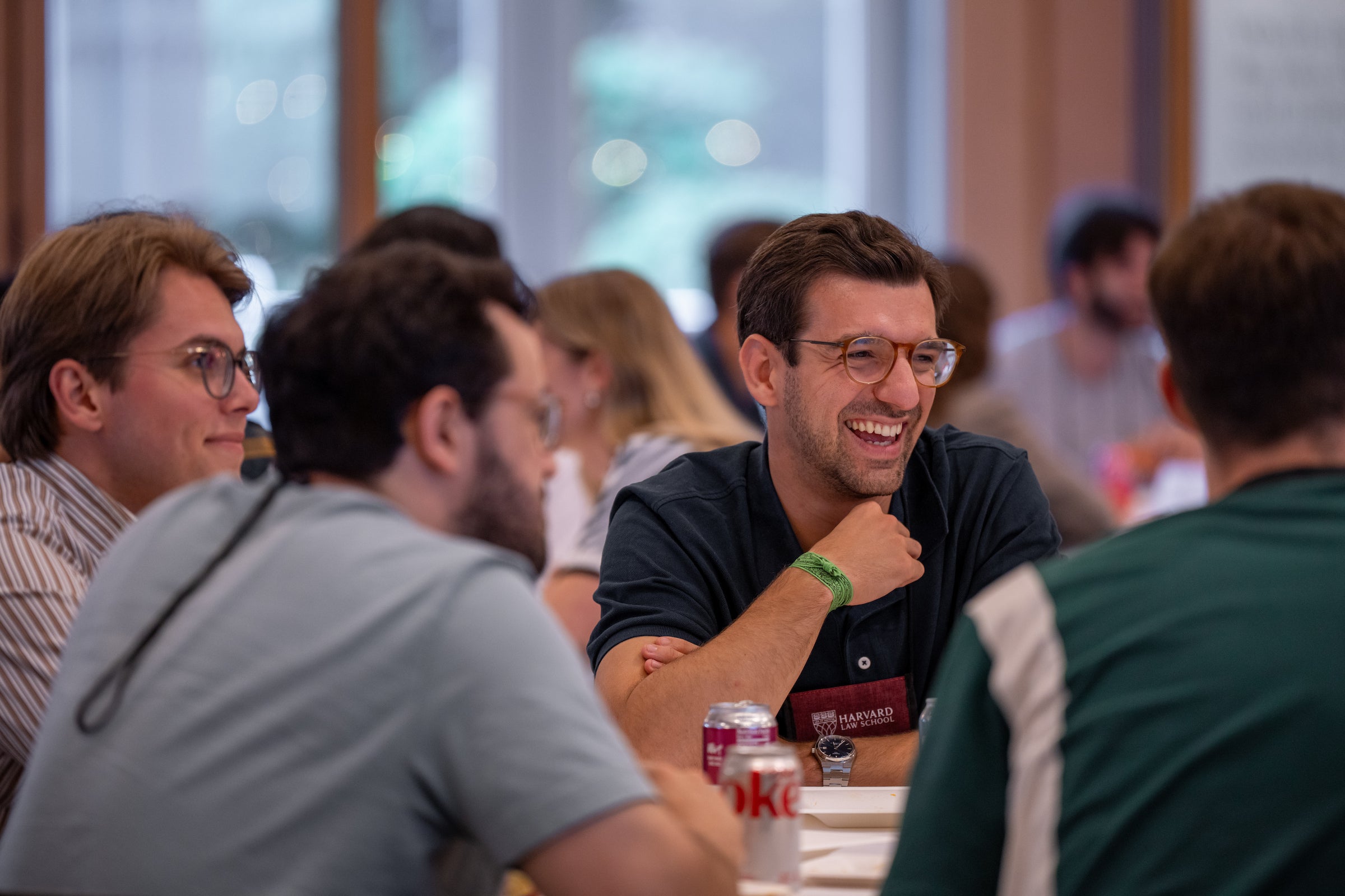 A group of students are gathered around a table, engaged in conversation, with one individual laughing while resting his chin on his hand.