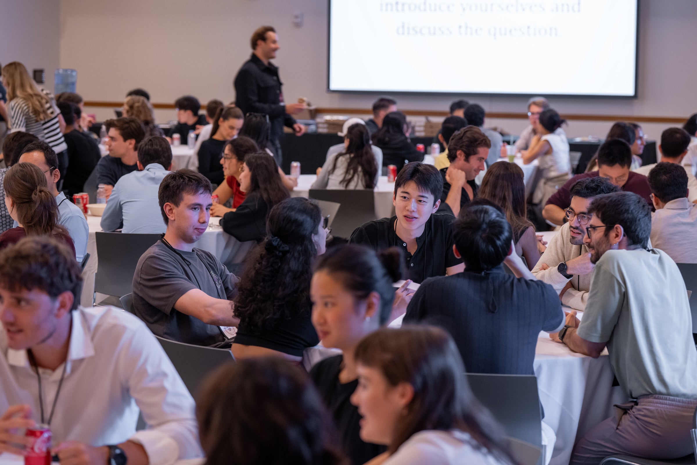 A group of students are seated at tables in a conference room, engaged in discussion.