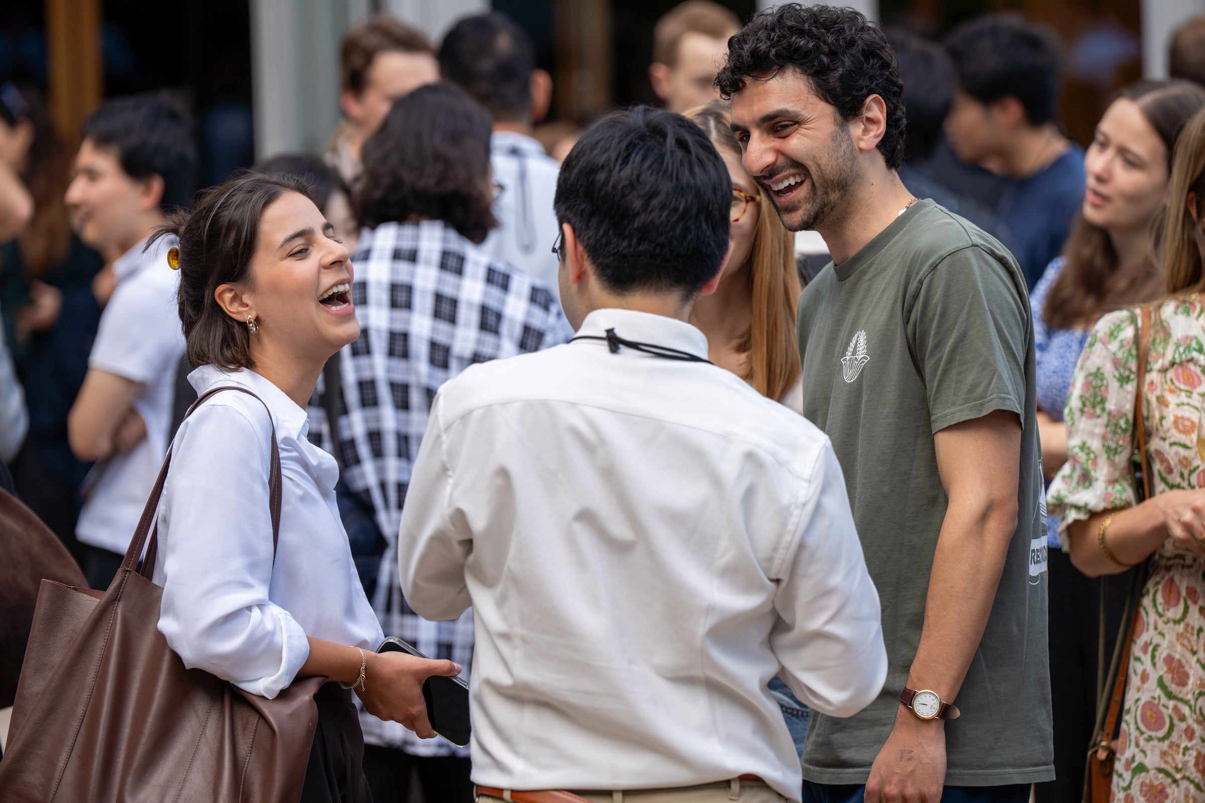 Three individuals laugh together in a lively outdoor gathering, surrounded by other people.