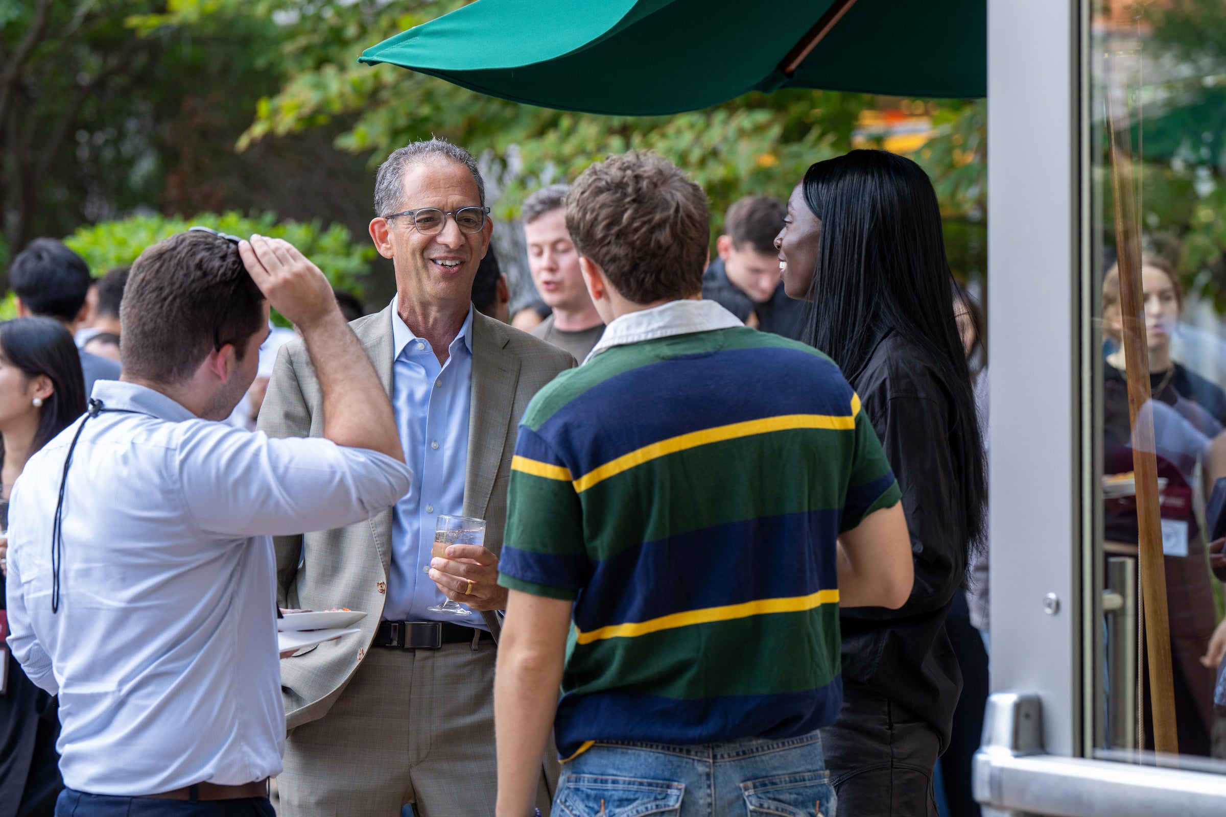 A group of students socialize and laugh with the dean outdoors under a green umbrella.
