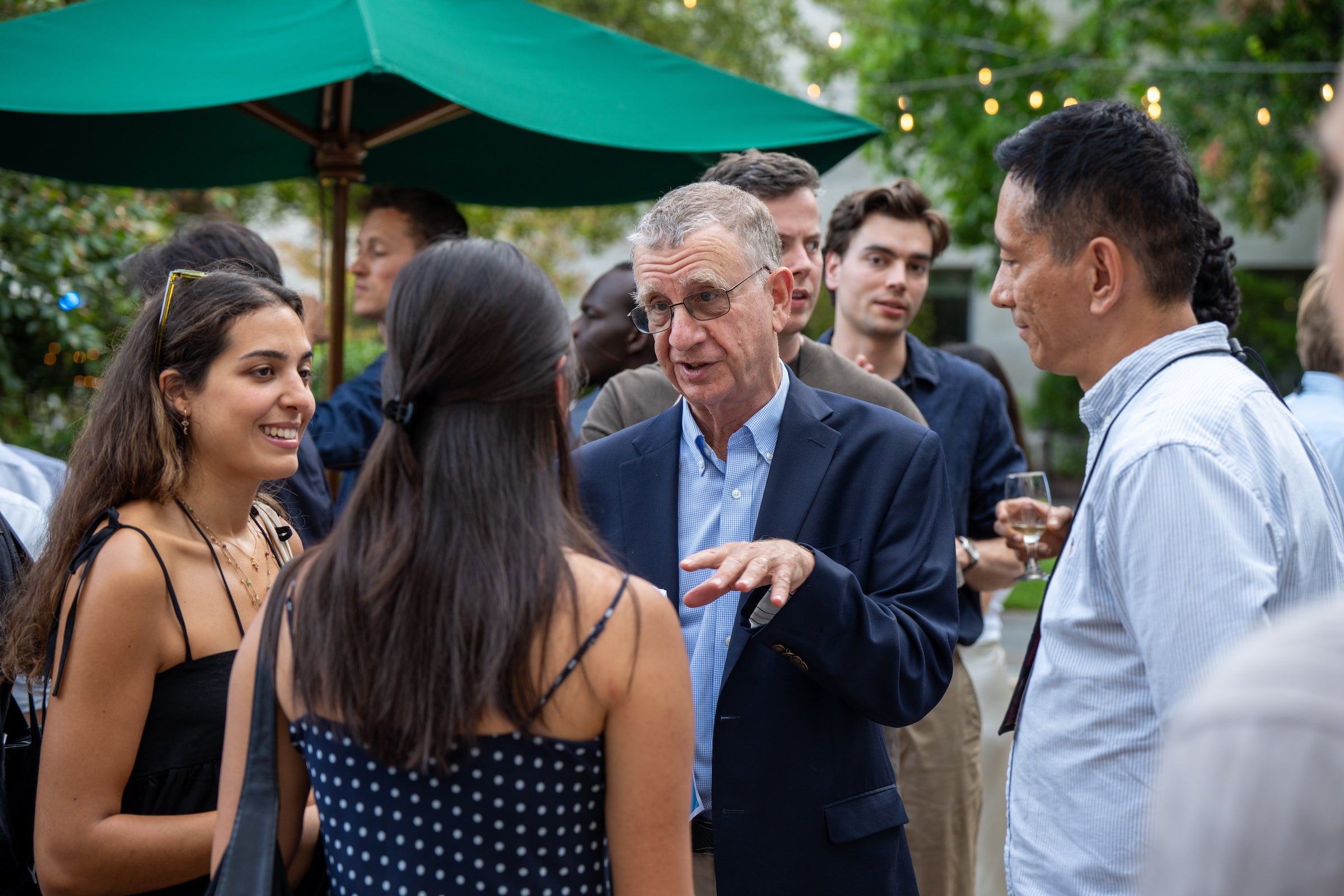 A group of students socialize with a professor outdoors under a green umbrella.