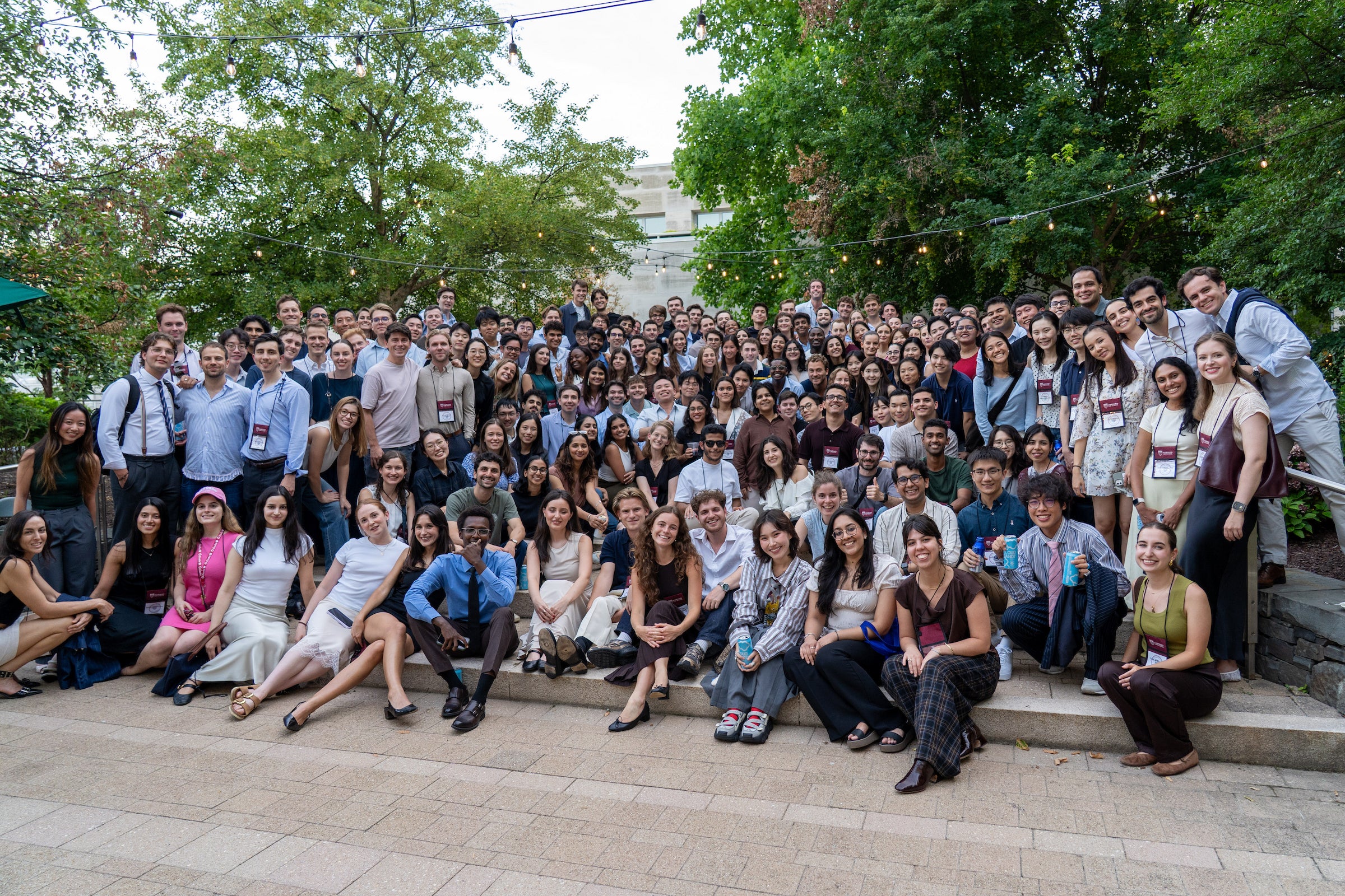A large group of people gather outdoors on stone steps for a group photo under string lights and trees.