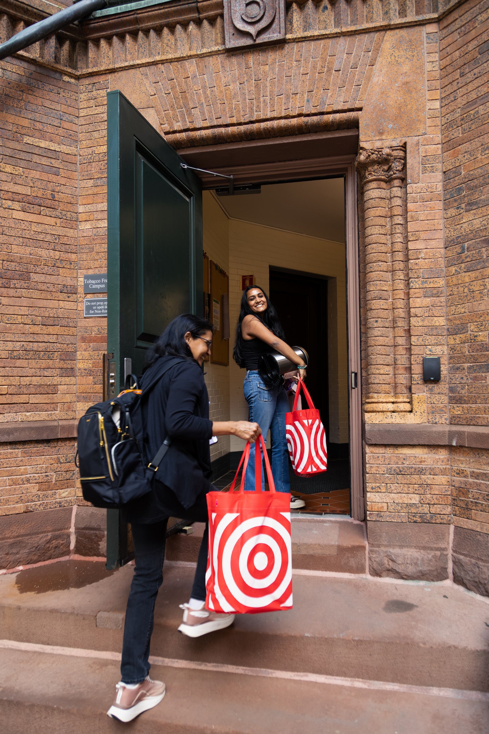 Two individuals enter a brick building, each carrying a red and white shopping bag during move-in.