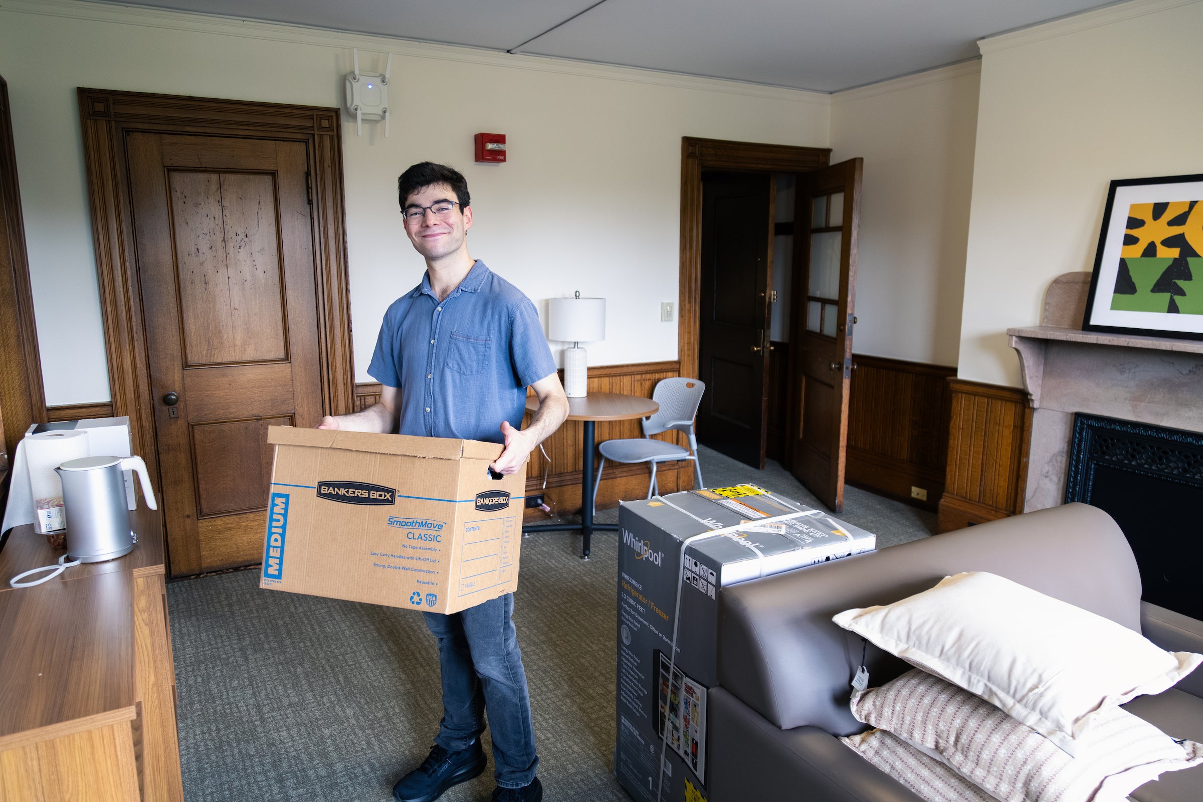 A student carries a moving box and smiles at the camera while moving into a residence hall.