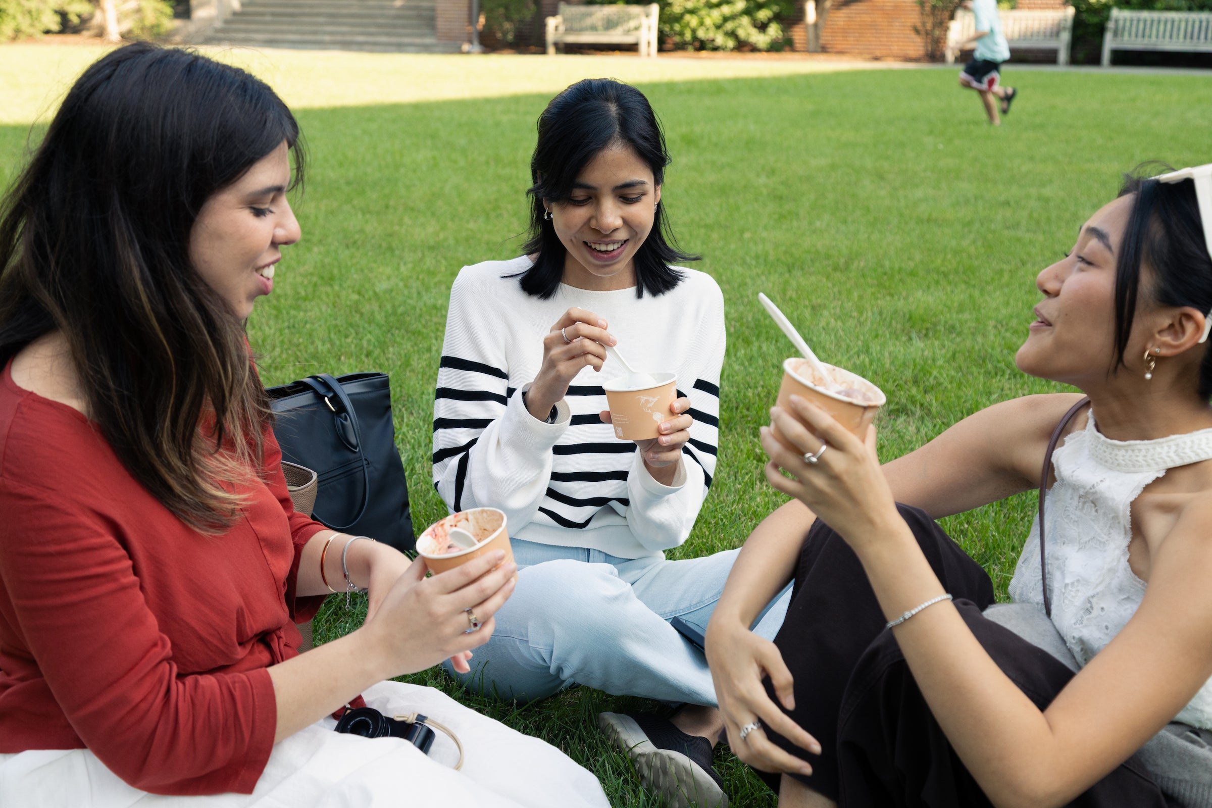 Three students enjoy eating ice cream while sitting on a grassy lawn at an outdoor event.