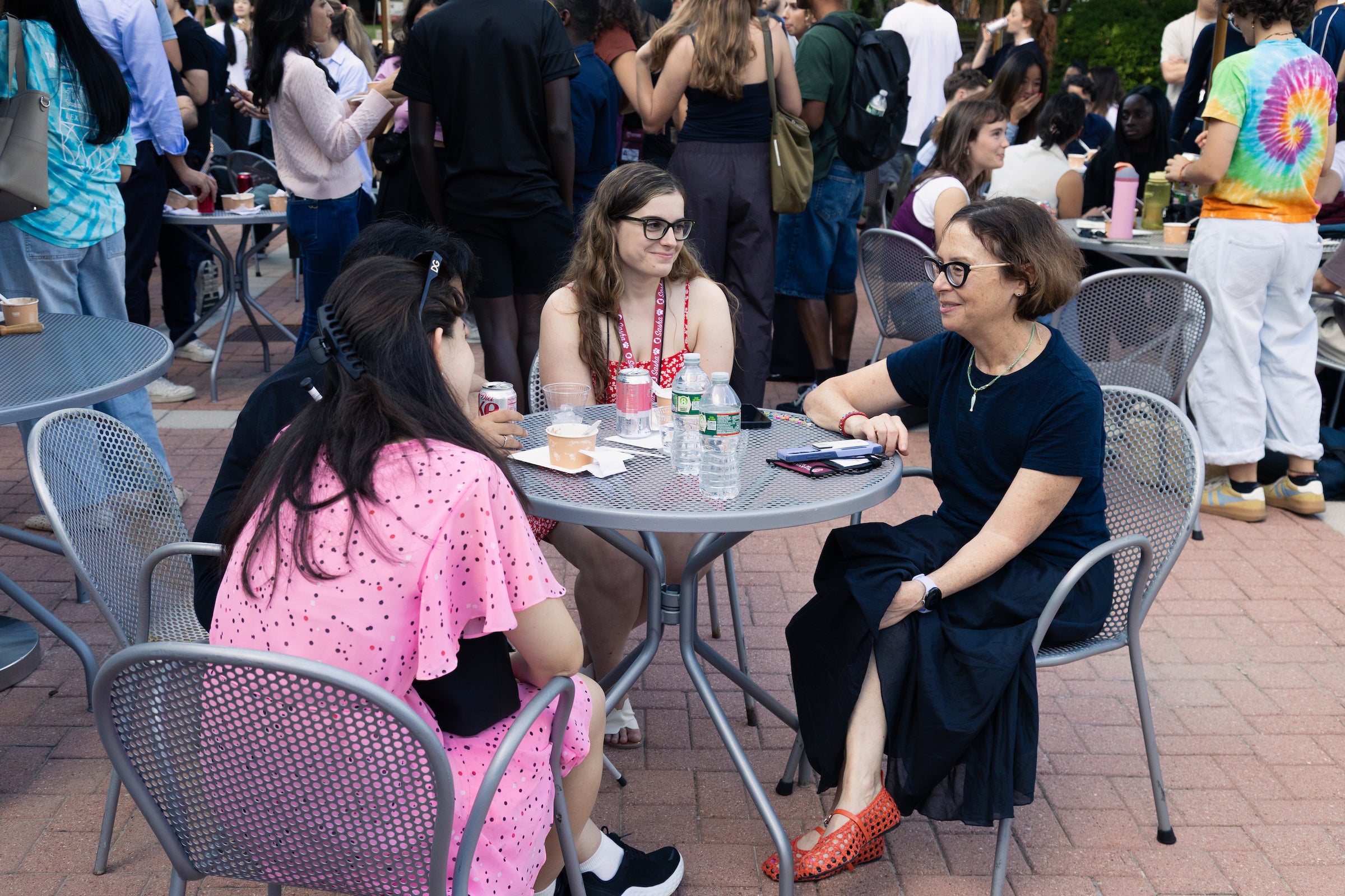 Three individuals engage in conversation at an outdoor table at a social gathering surrounded by people.
