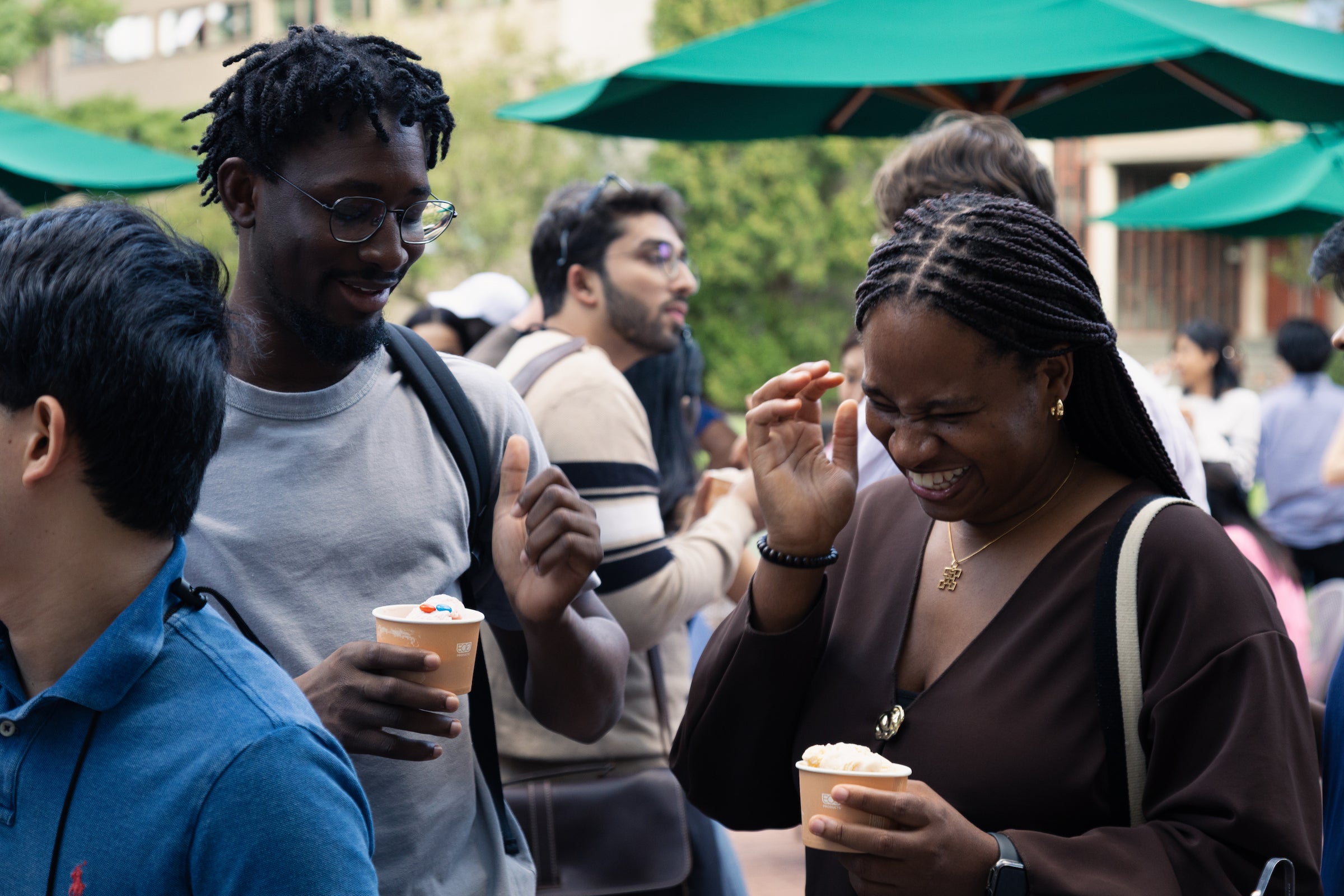 Two students laugh and enjoy ice cream together at an outdoor gathering under green umbrellas.