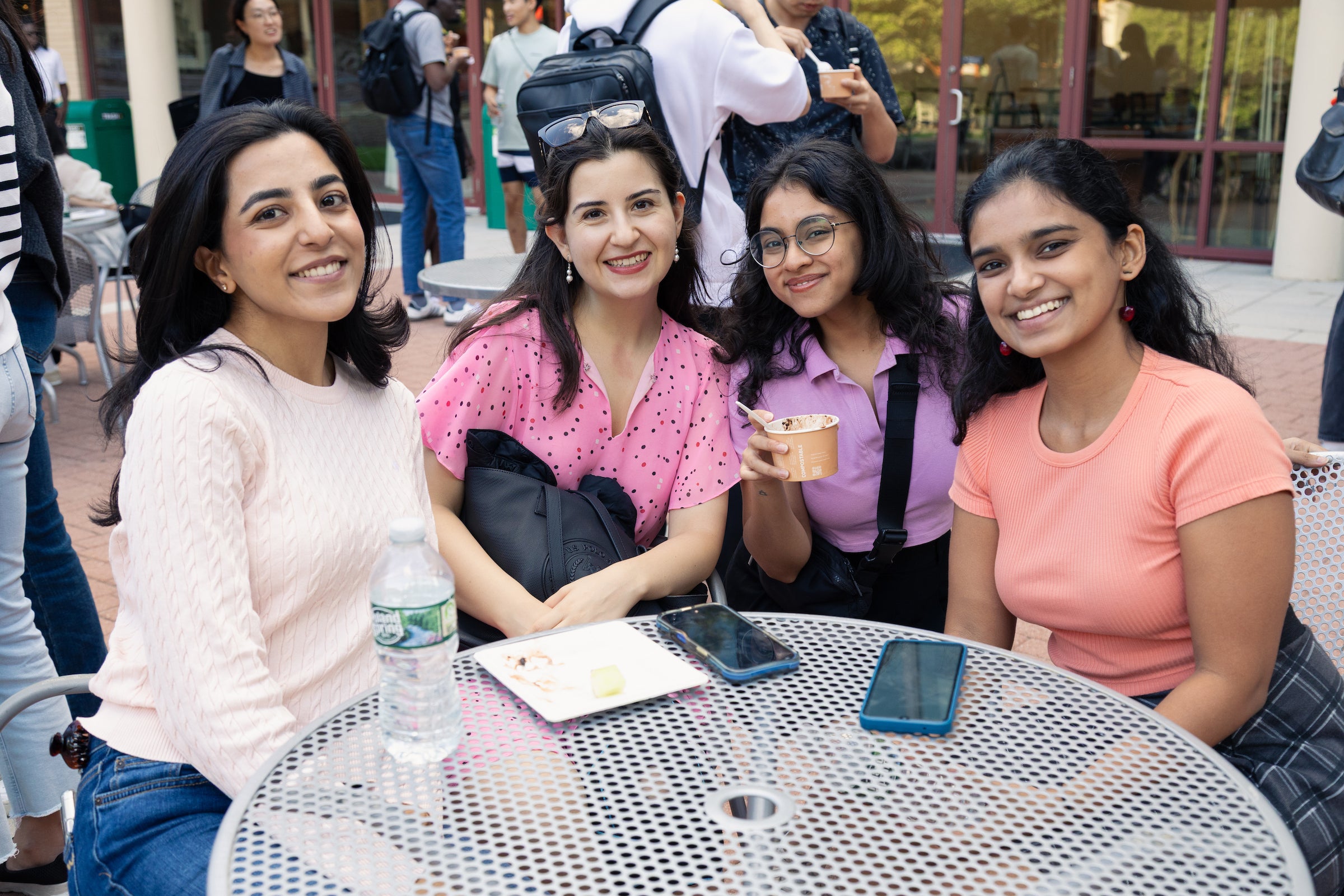 Four students sit together at an outdoor table, smiling for the camera, with one holding up a cup of ice cream.
