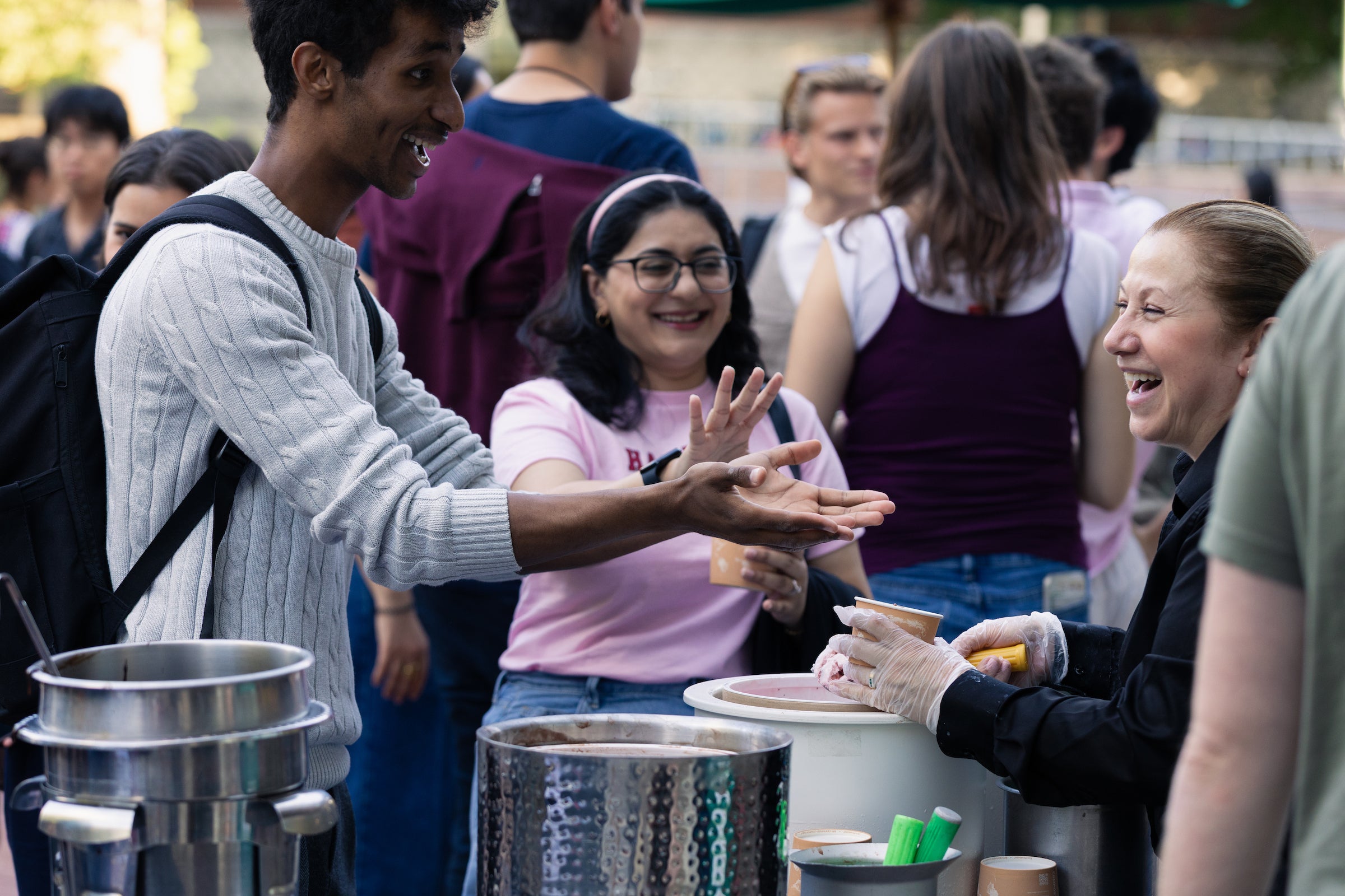 A staff member laughs as she serves two students ice cream at an outdoor event.