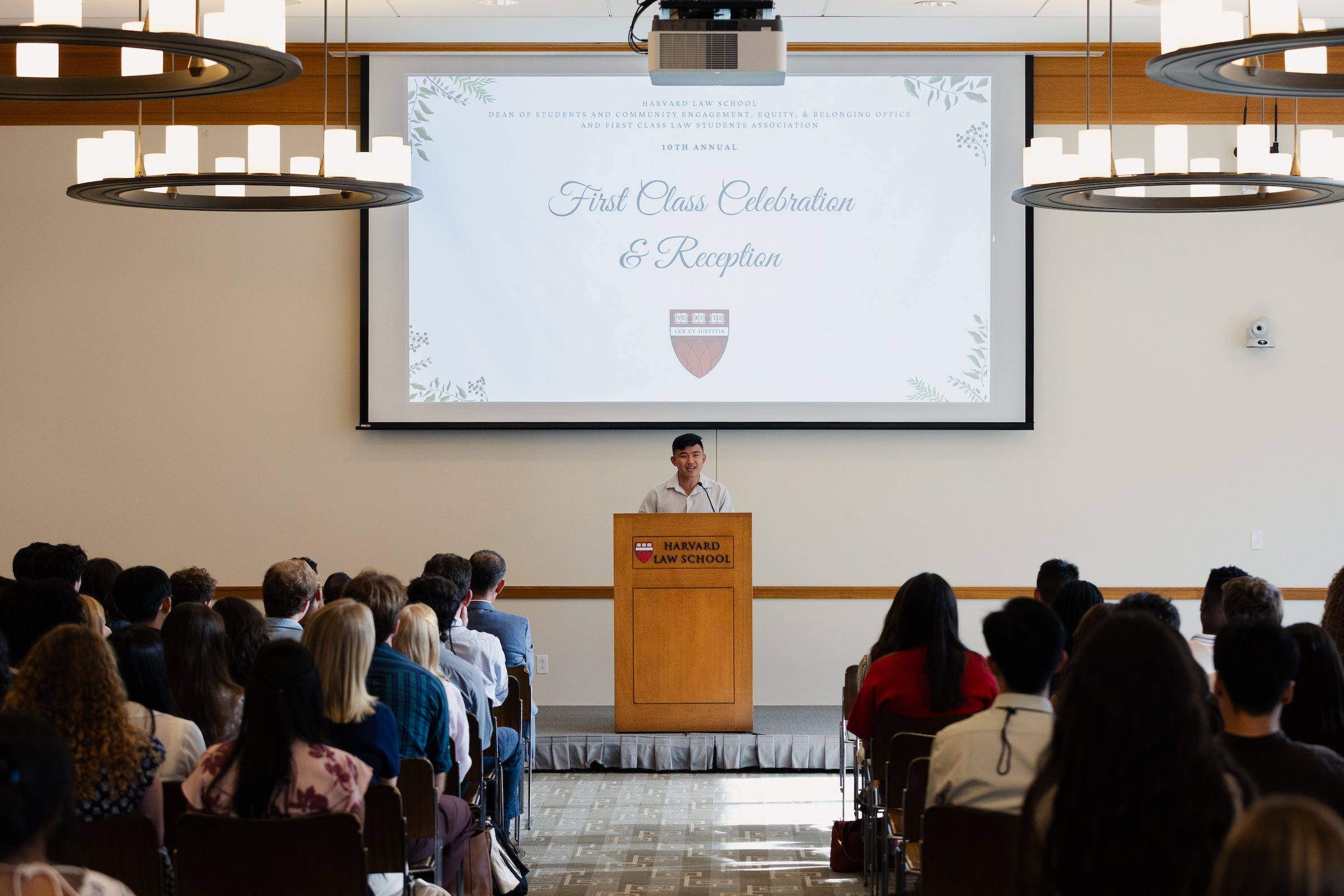 An individual speaks to an audience from a podium in front of a screen that reads 