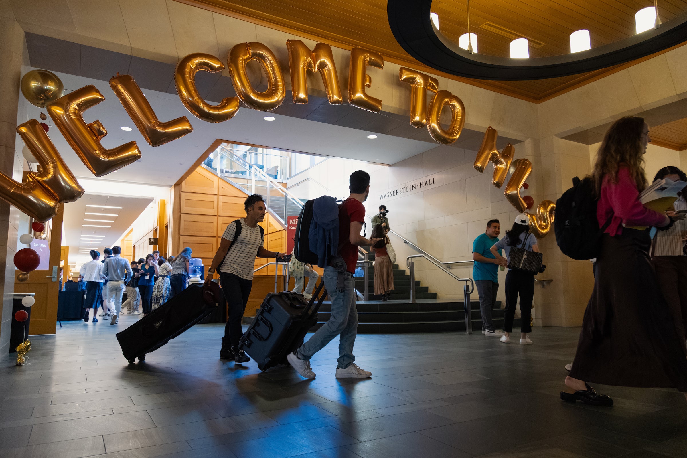 Students with luggage walk through a brightly lit hall adorned with large gold 
