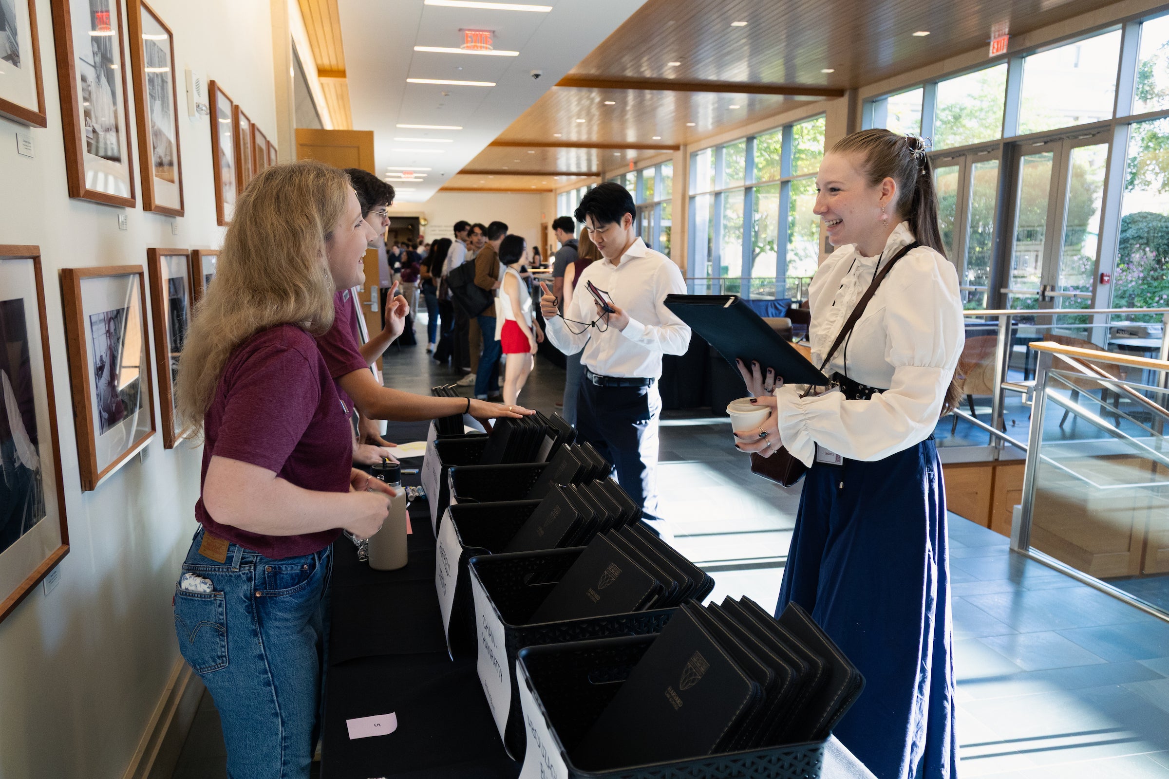 Students check in a table with black folders, engaging in conversation in a brightly lit, modern hallway lined with framed photographs.