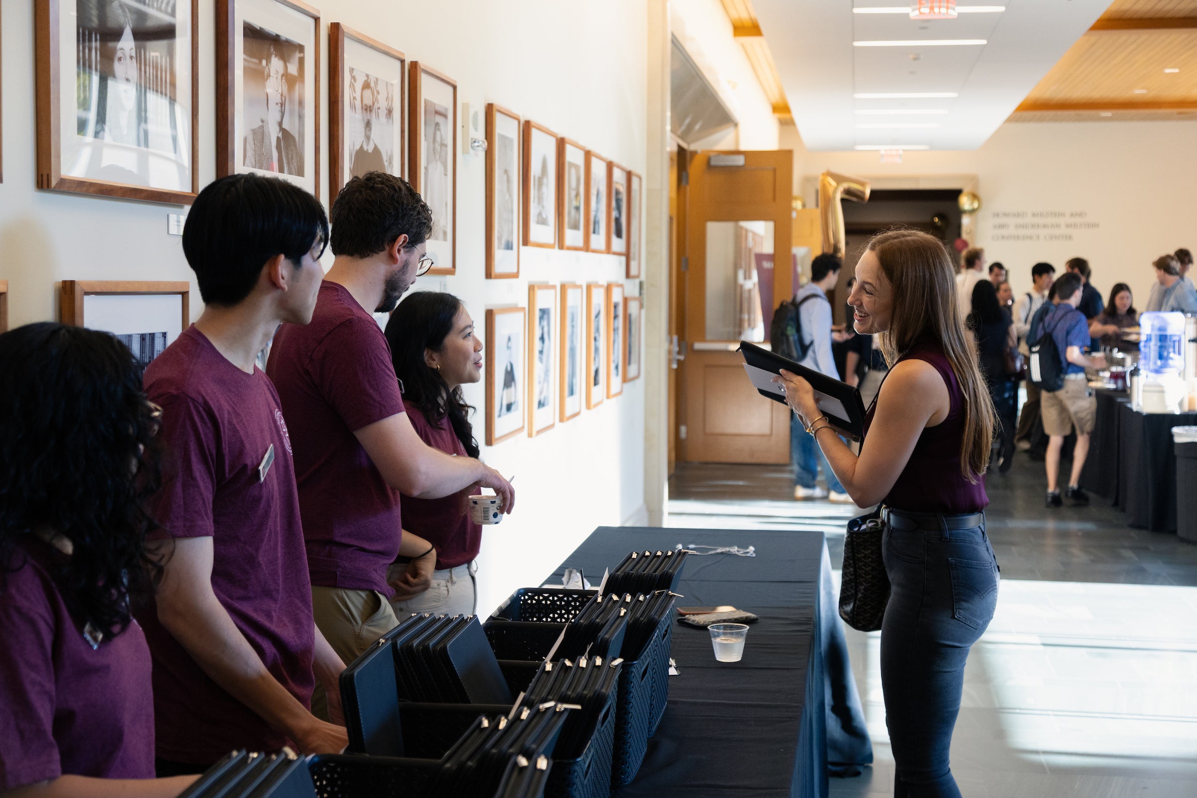 Students check in a table with black folders, engaging in conversation in a brightly lit, modern hallway lined with framed photographs.