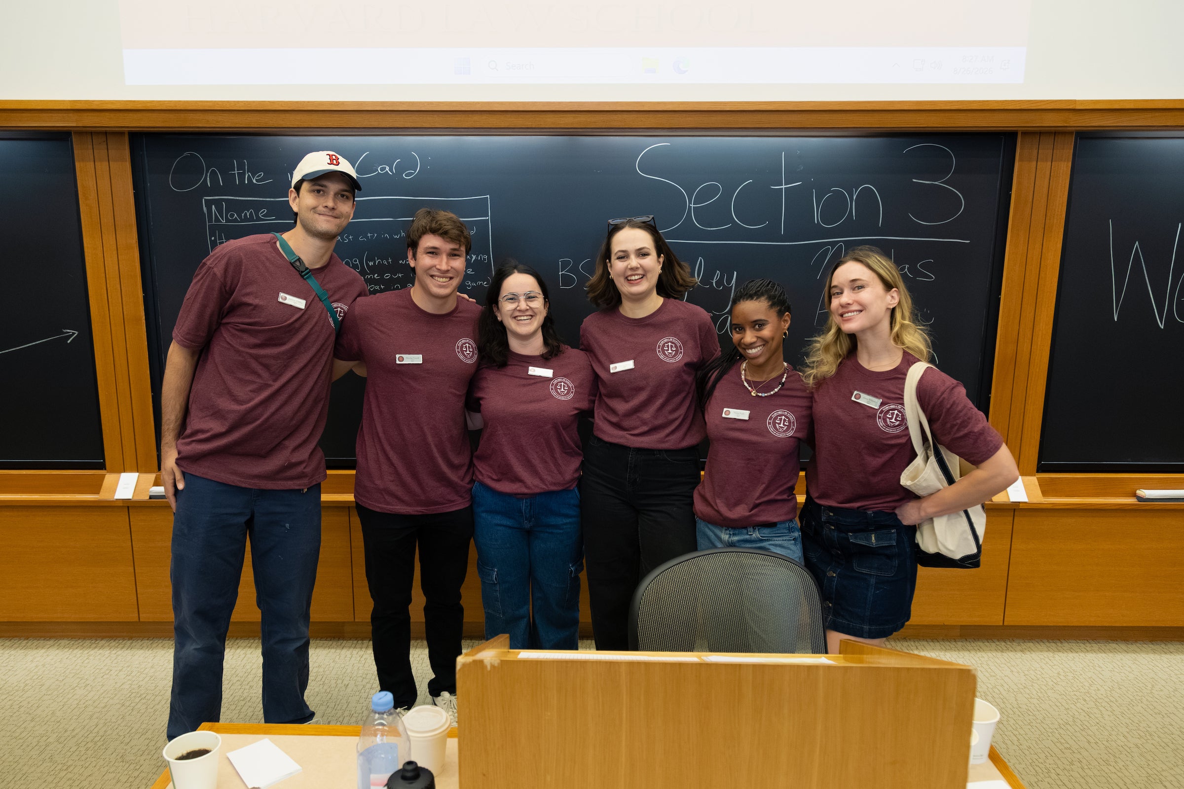 Six students smile for a group photo in front of a blackboard that has 