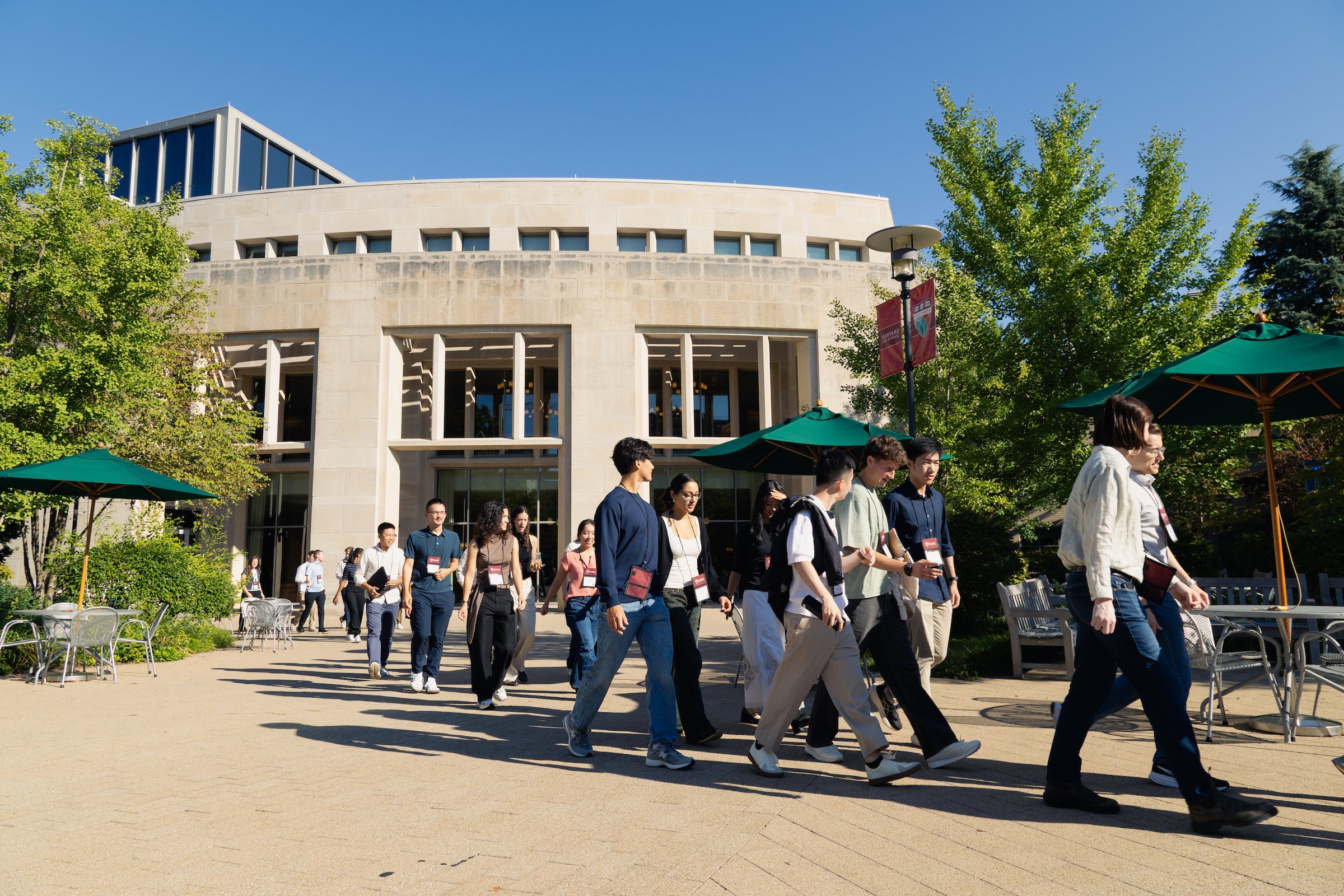 A group of students walk across a paved courtyard in front of a modern building on a sunny day, surrounded by green trees and outdoor seating with green umbrellas.