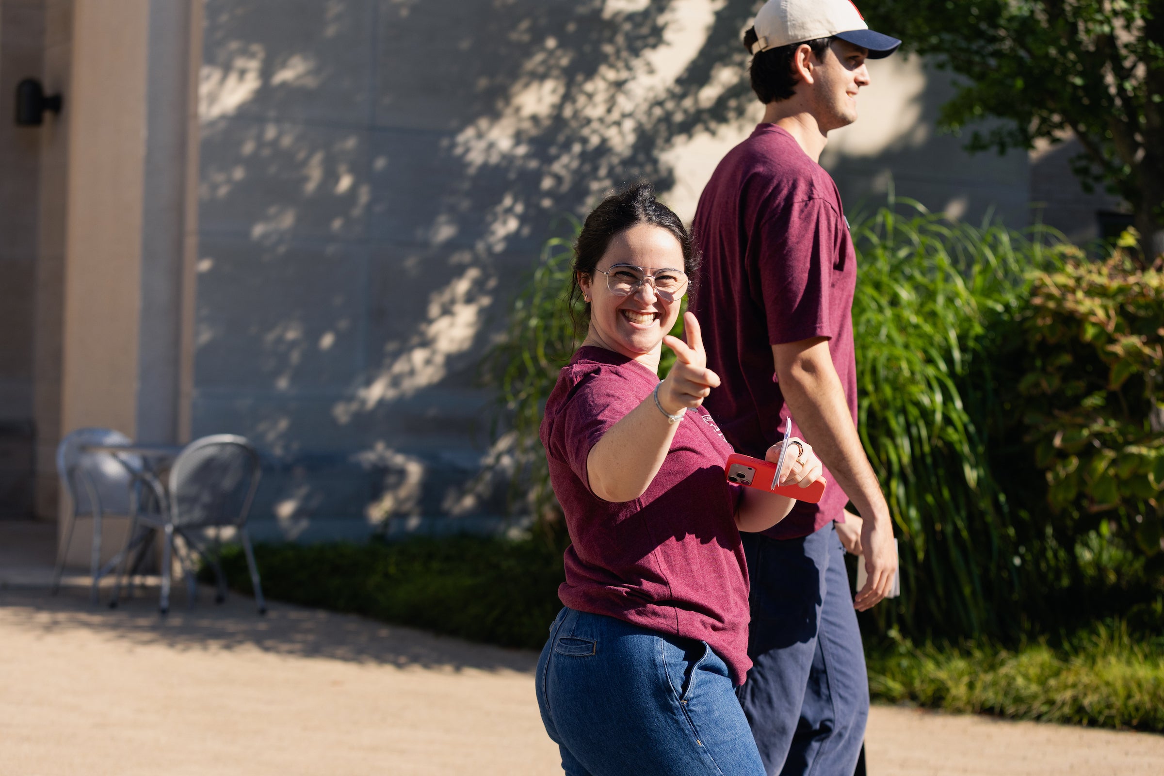 Two people in maroon shirts walk outdoors, with one smiling and pointing toward the camera.