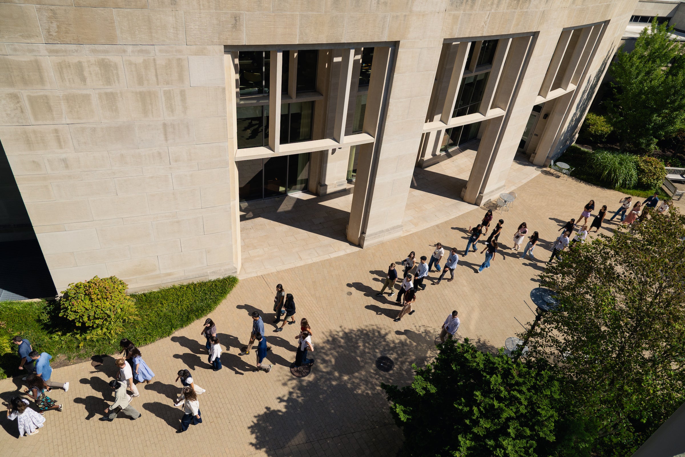 A group of students walk across a paved courtyard beside a large stone building on a sunny day, taken from a high angle.