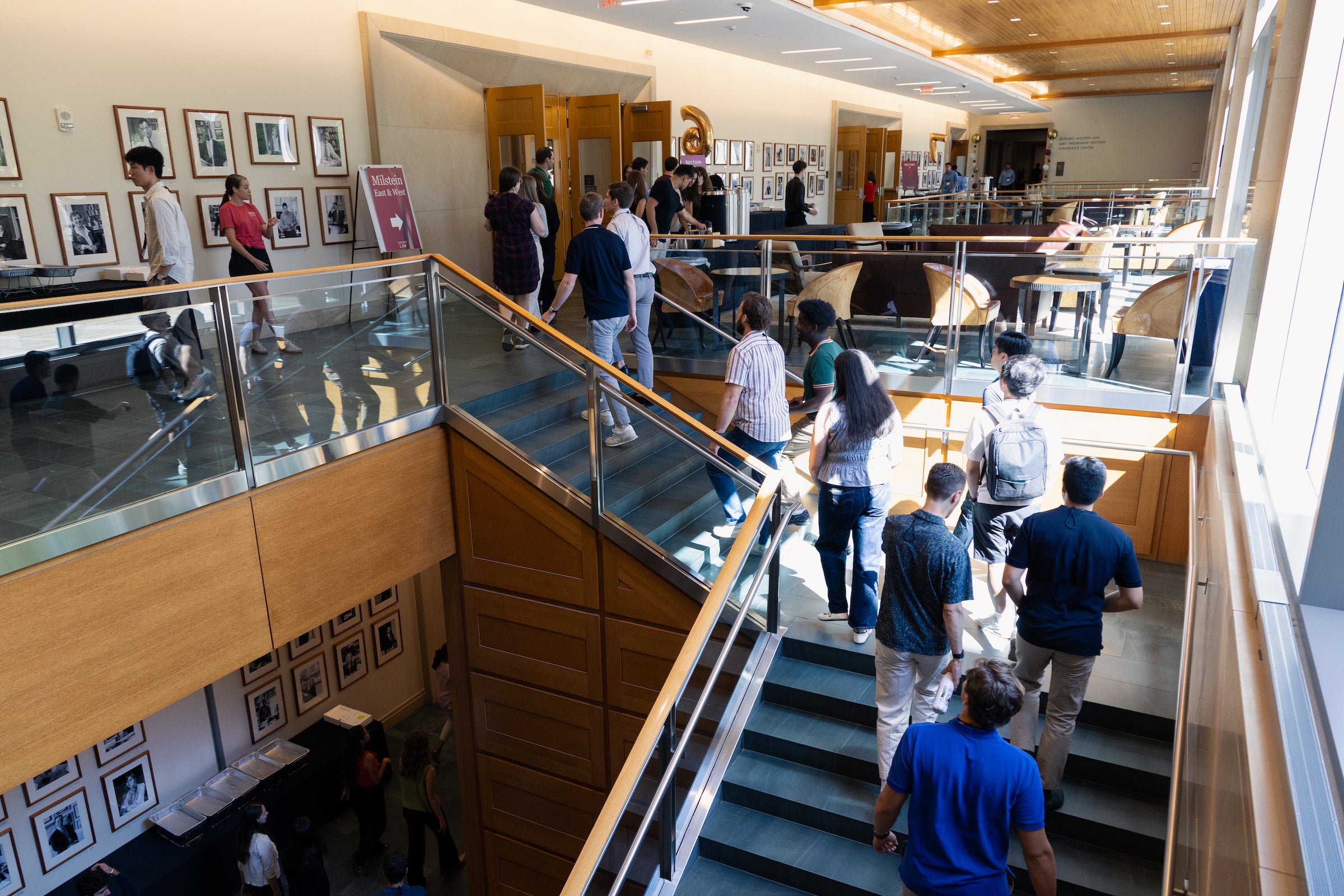 A group of students ascend a modern staircase in a well-lit, multi-level building with framed photographs on the walls and glass railings.