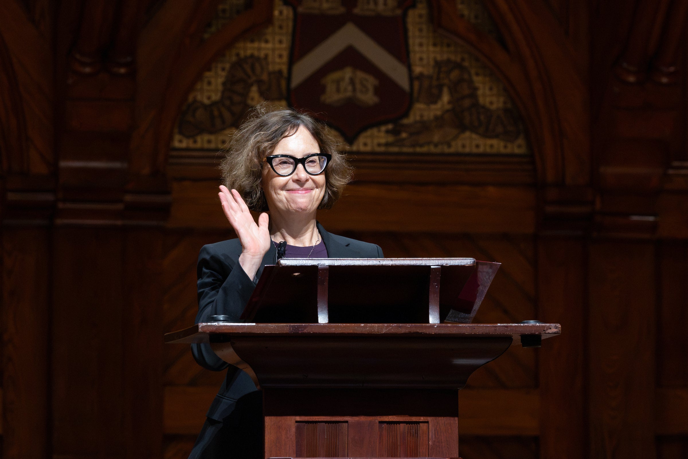 A speaker smiles and waves at an audience from a wood podium.