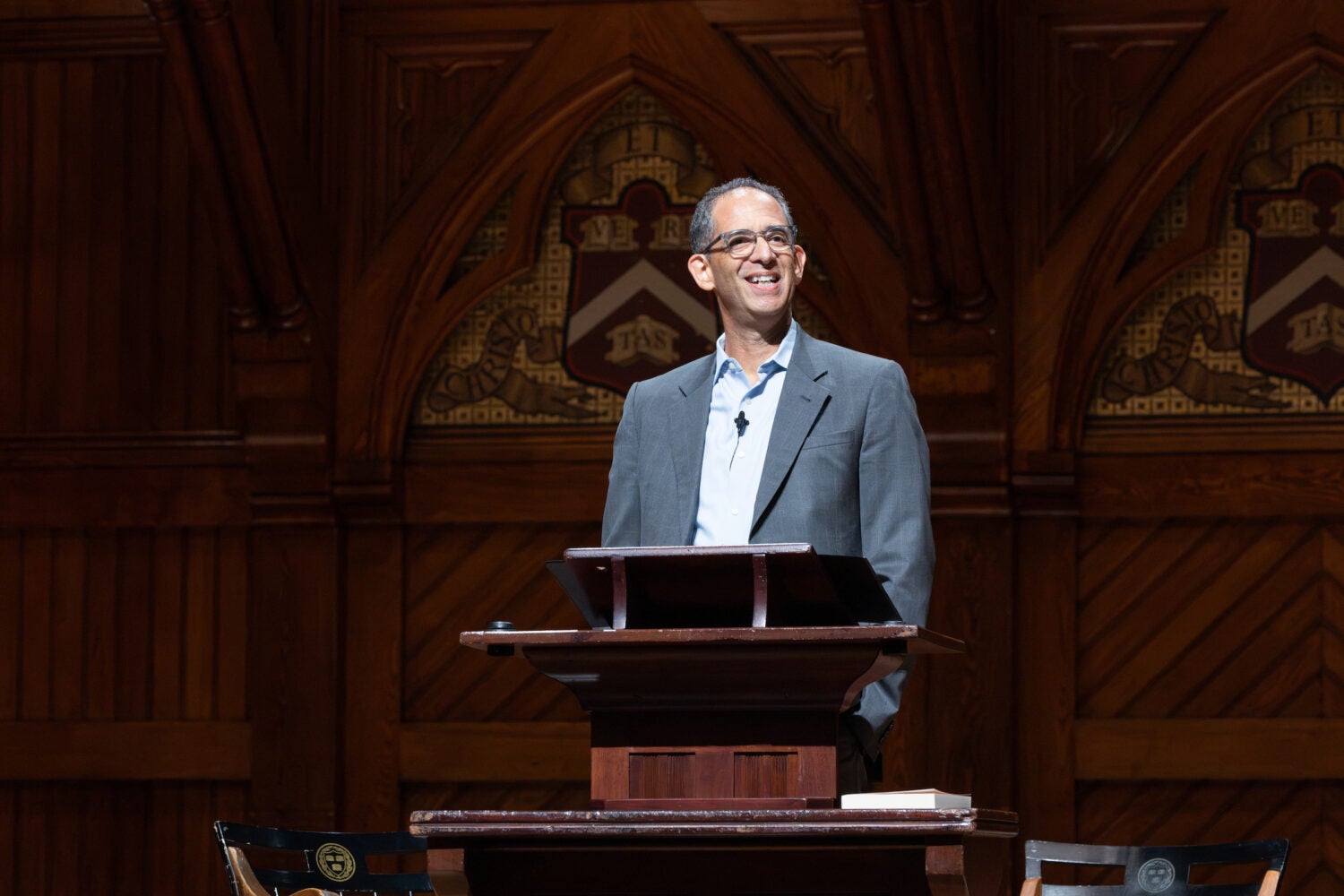 Dean Goldberg smiles at an audience while standing at a wooden podium in front of ornate wooden paneling with decorative shields.
