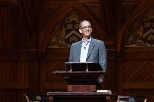 Dean Goldberg smiles at an audience while standing at a wooden podium in front of ornate wooden paneling with decorative shields.