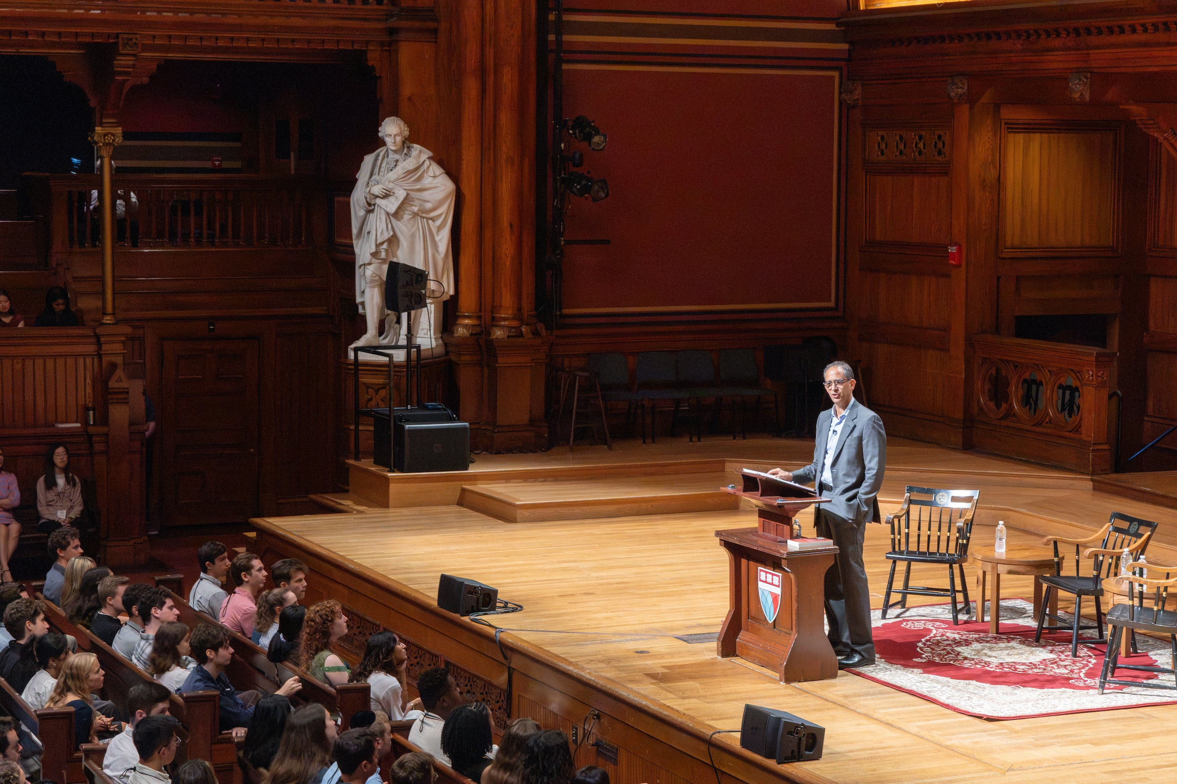 Dean Goldberg stands at a podium on a wooden stage in a historic theatre, addressing an audience seated in rows, with a large statue visible in the background.