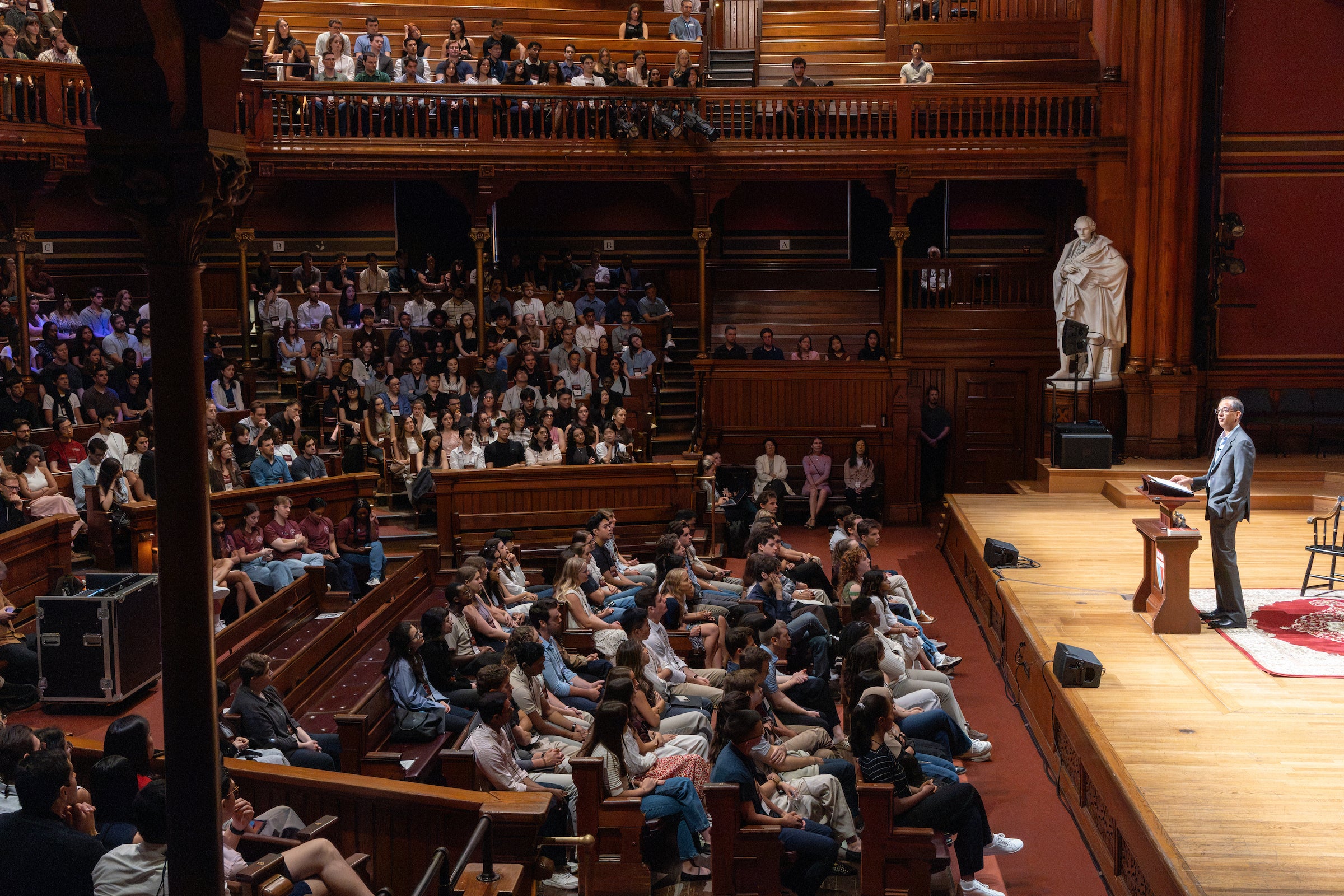 Dean Goldberg addresses students from a podium in a historic, wood-paneled theatre with a statue of a man in the background.