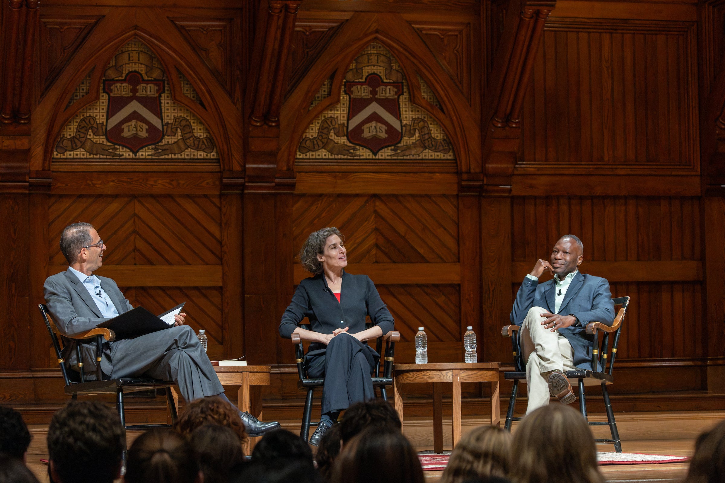 Dean Goldberg and two professors engage in a discussion on a wooden stage, seated in chairs with ornate wooden paneling and decorative shields behind them.