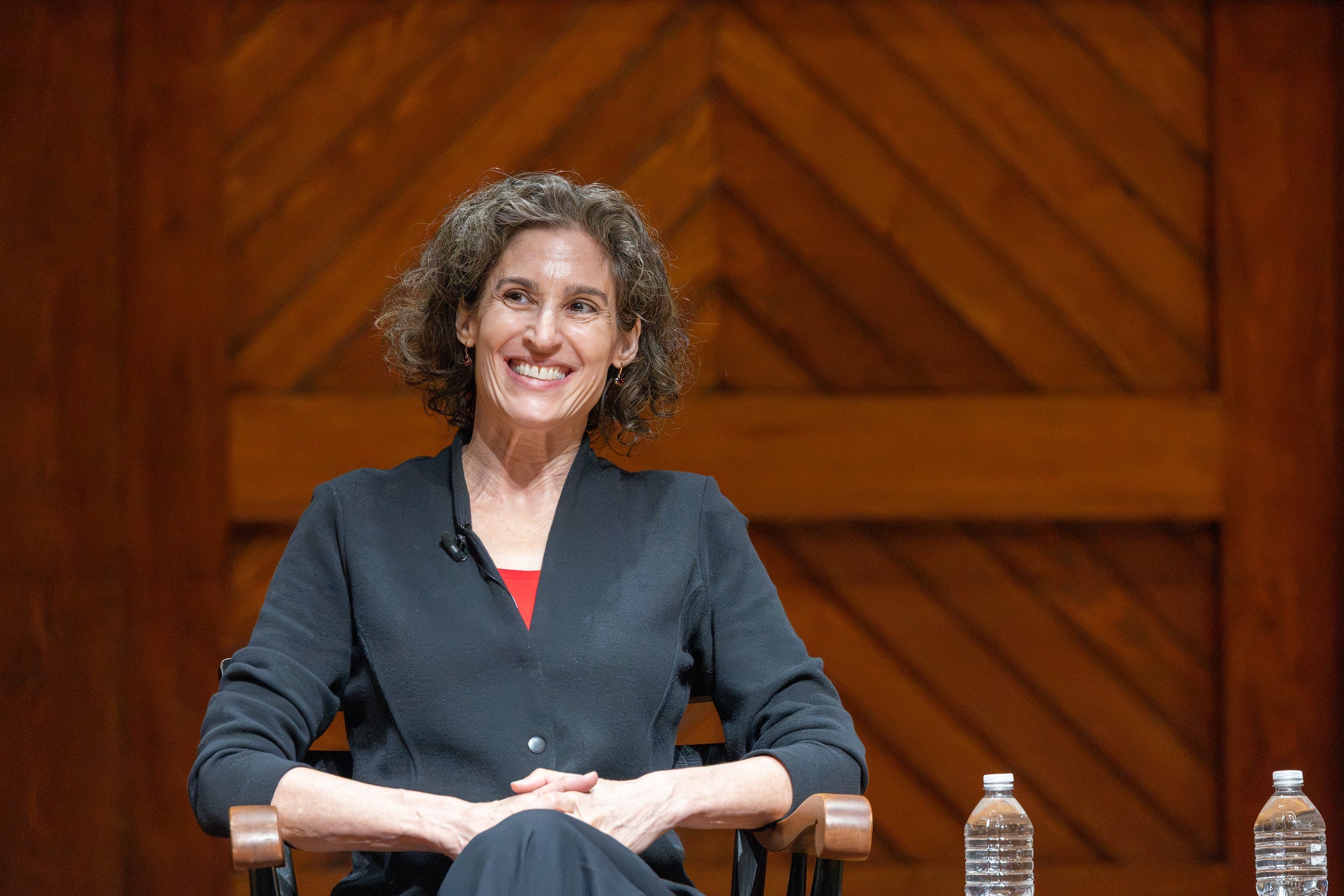 A professor smiles at an audience while seated on a stage.