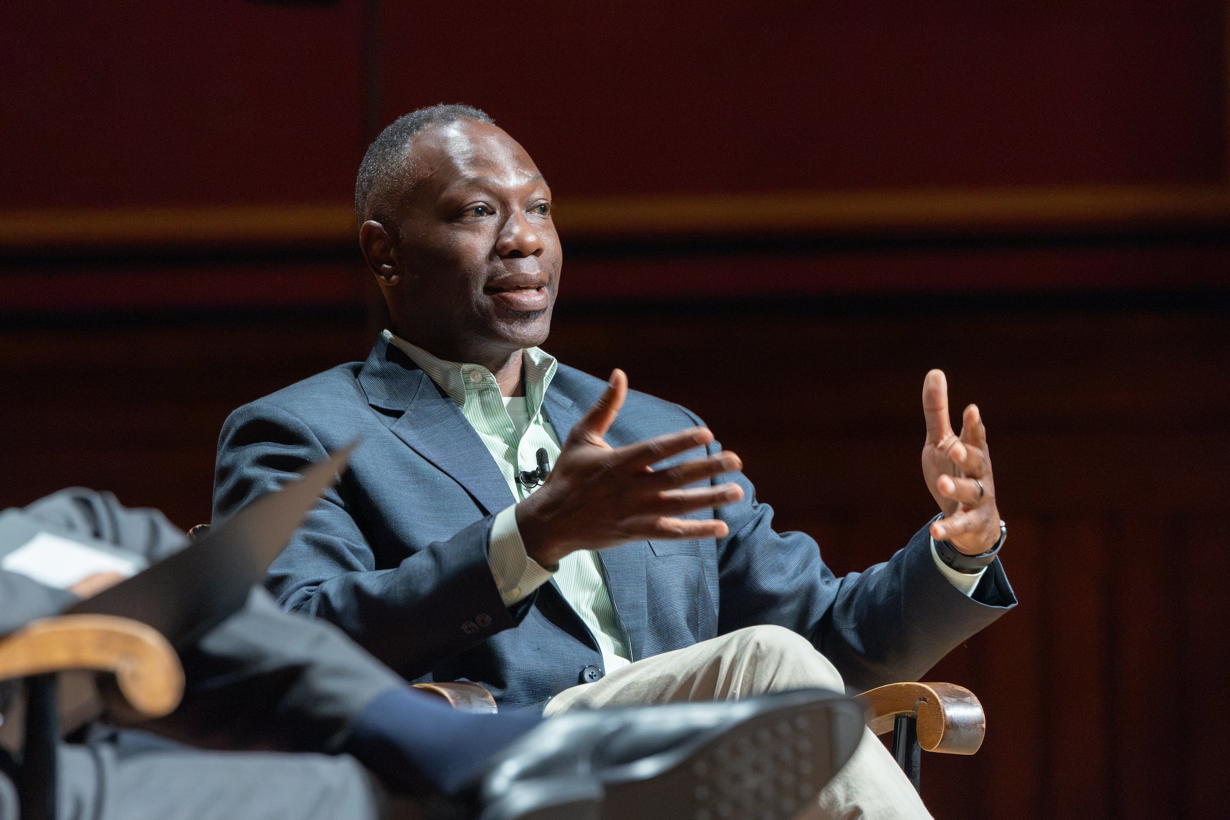 A professor speaks to an audience while seated on a stage.