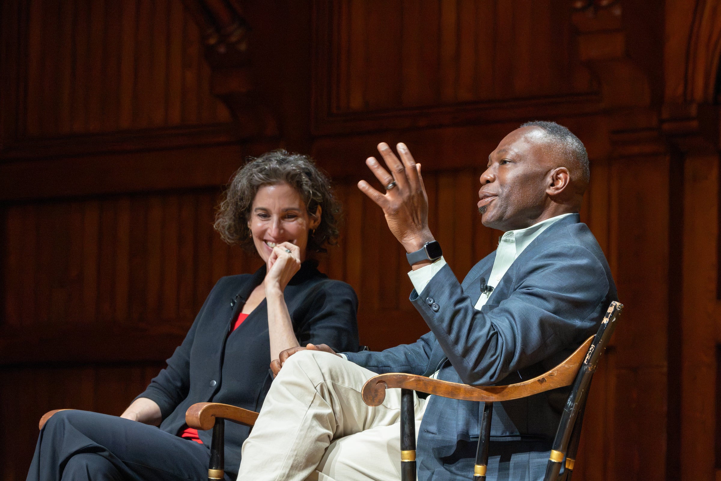 Two professors engage in a conversation during a seated discussion, with one gesturing while speaking and one laughing.