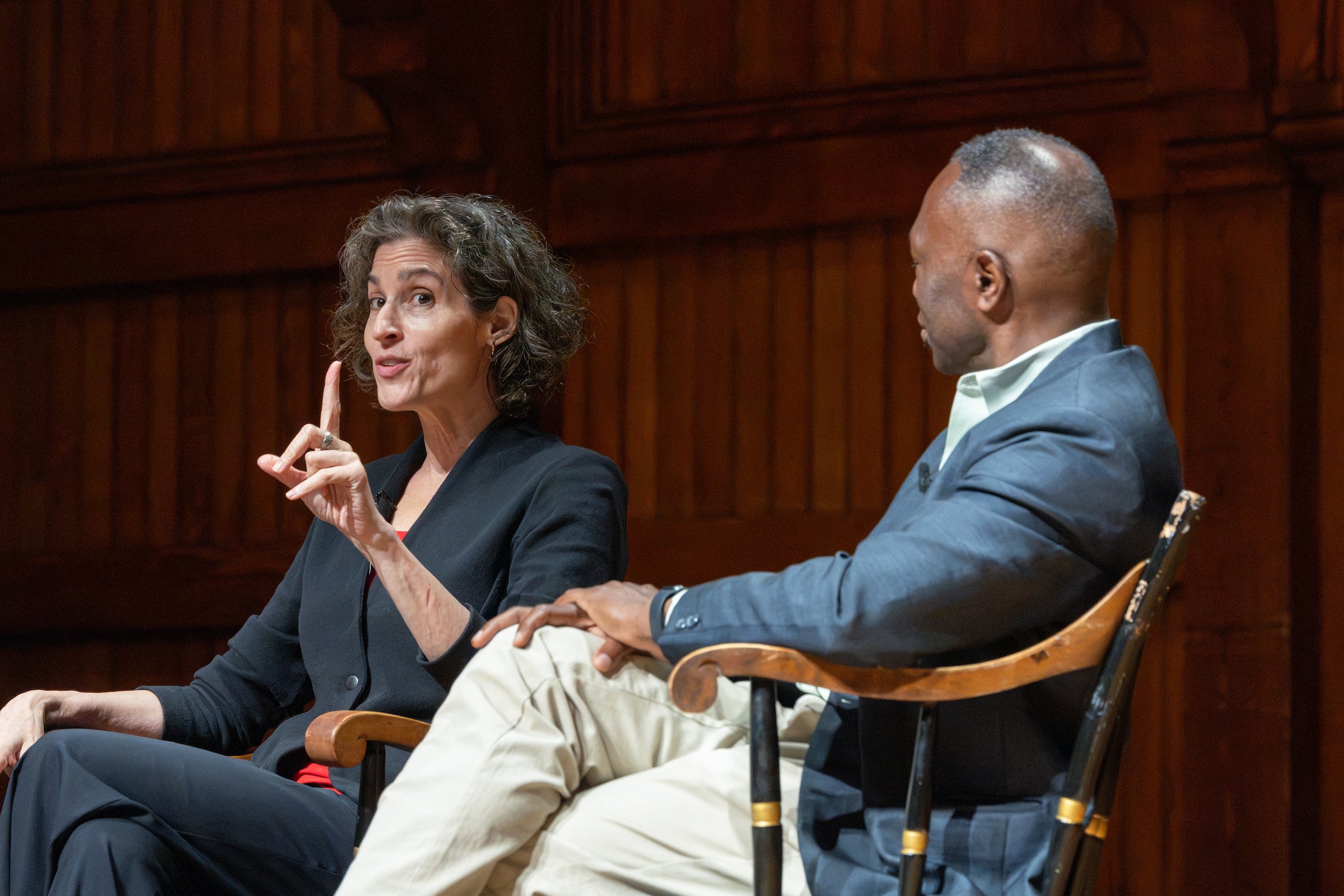 Two professors engage in a conversation during a seated discussion, with one gesturing while speaking.