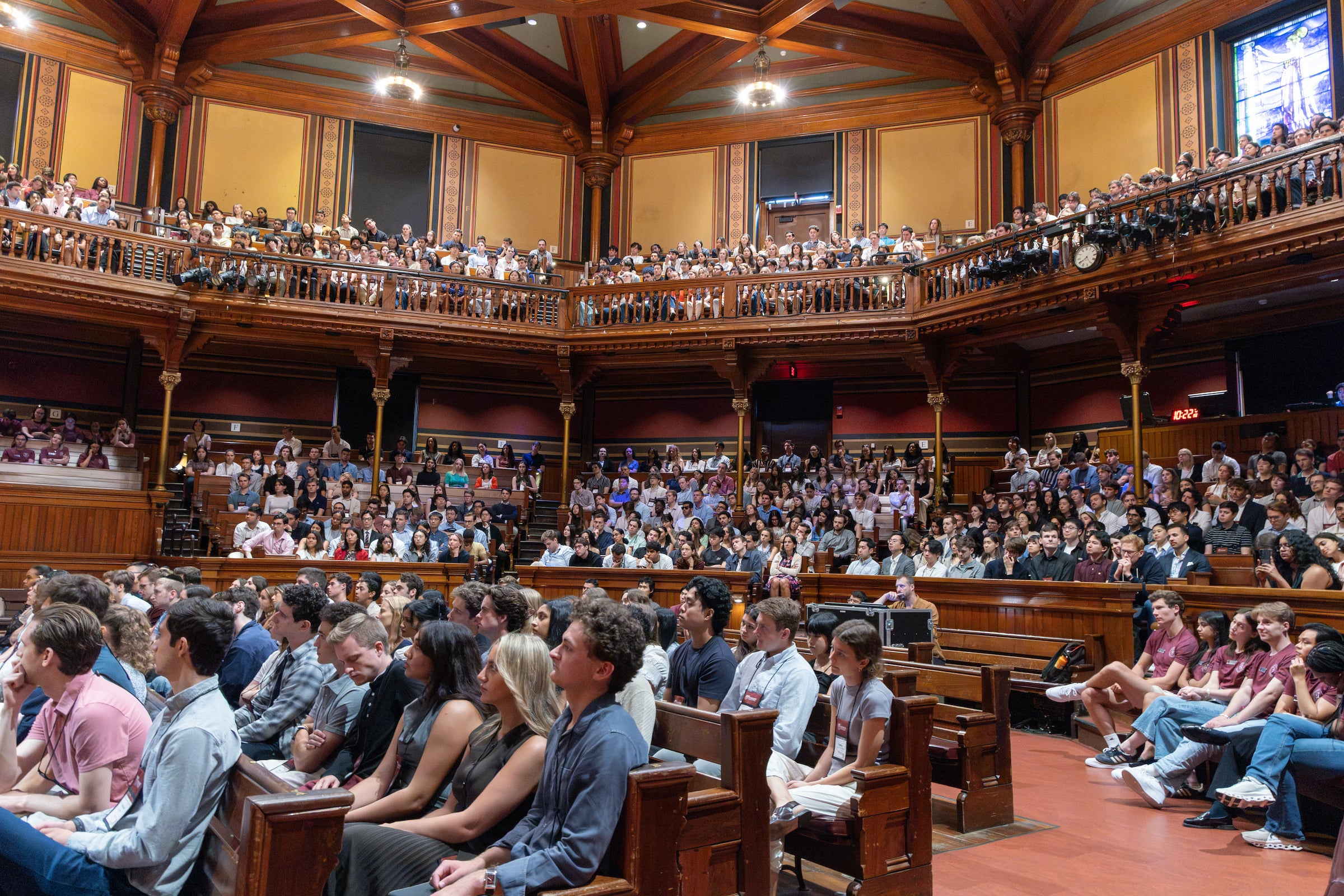 A large audience fills a grand, wood-paneled auditorium with tiered seating, attentively watching the stage.