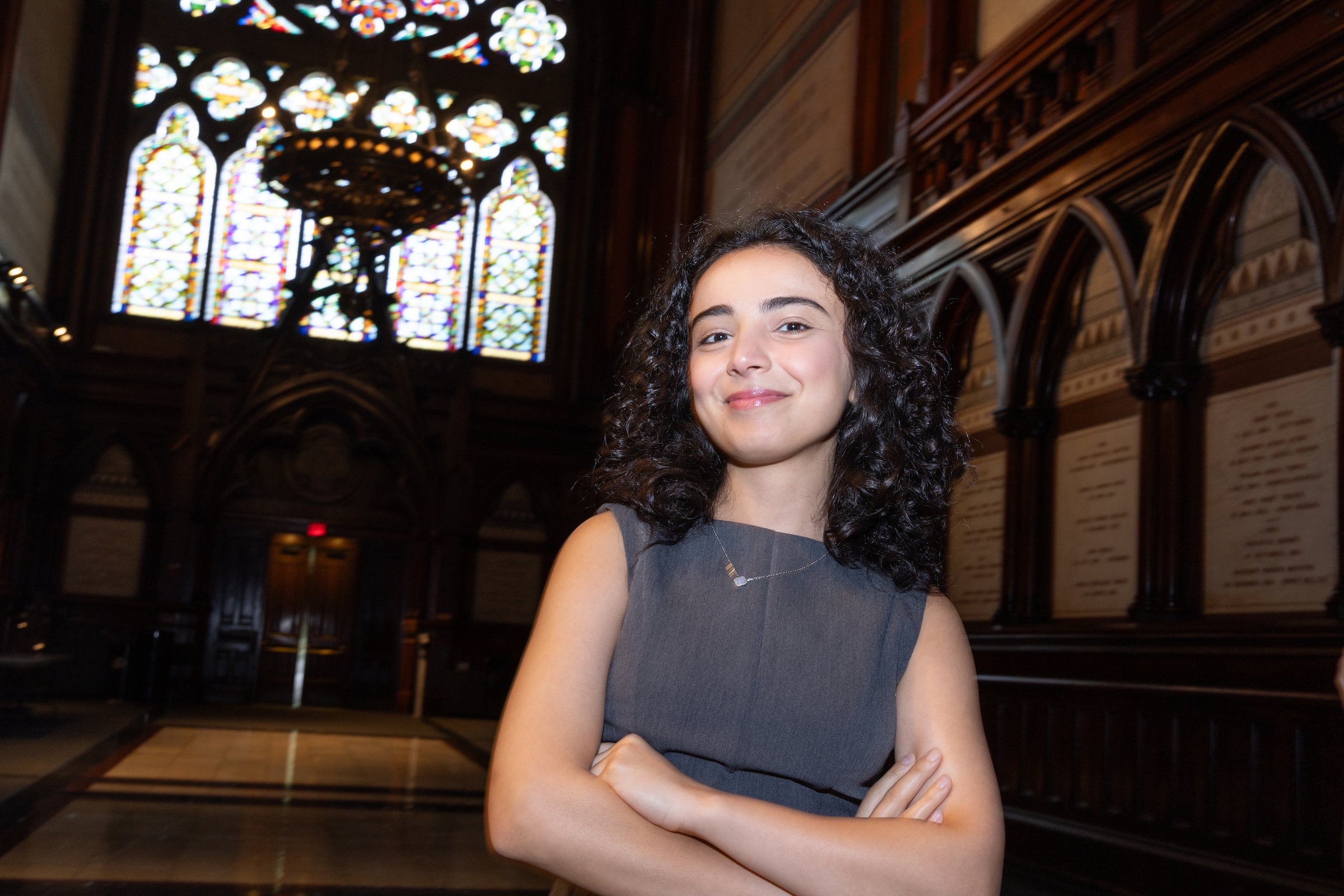A student stands with arms crossed in a grand hall, smiling beneath a large stained-glass window.